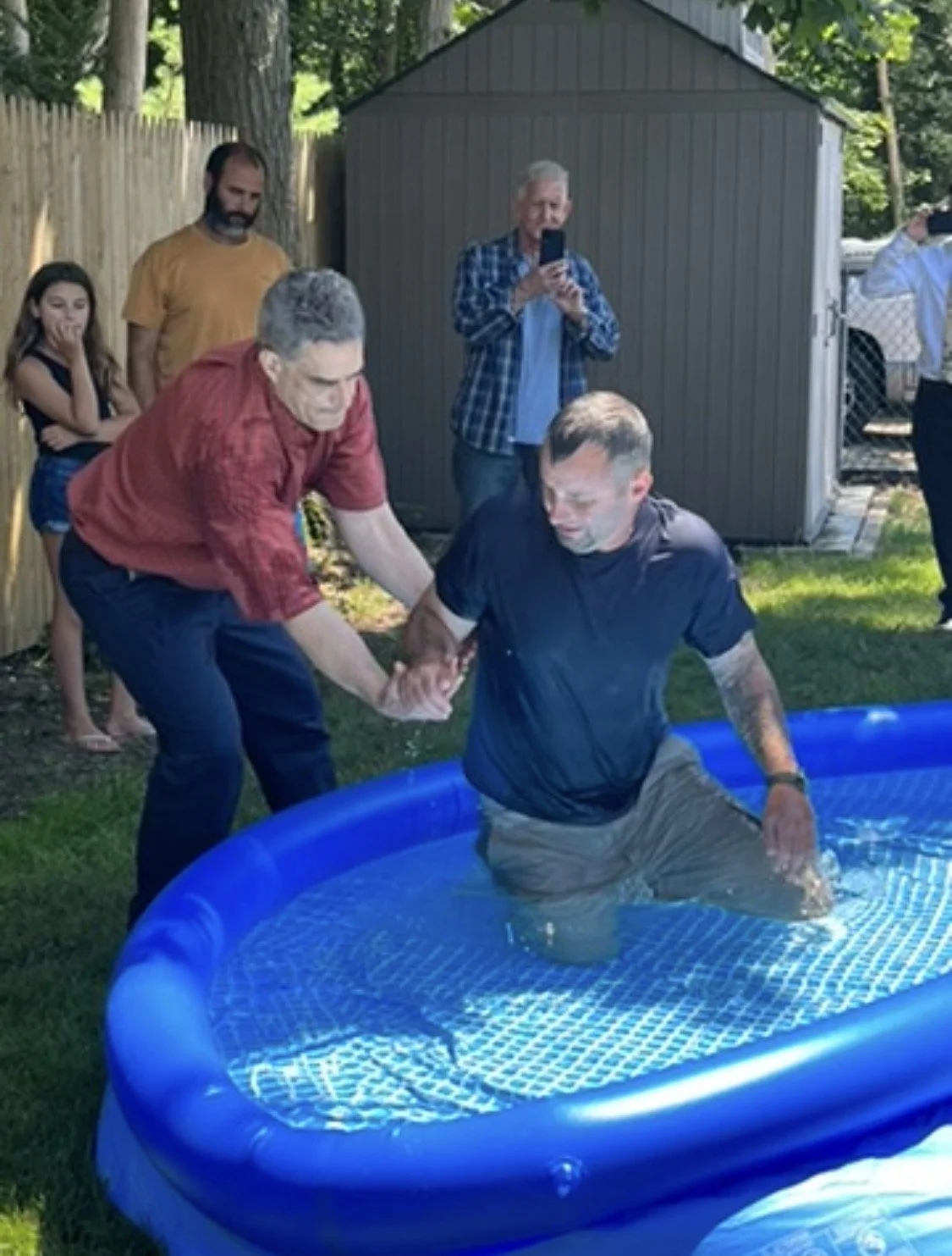 Two men are participating in a baptism ceremony in an inflatable pool, with one man holding the other's hand as he kneels in the water. Several onlookers, including a girl and adults, watch the event, with some taking photos.