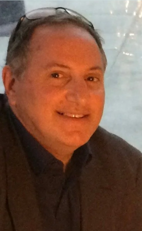 A smiling middle-aged man with glasses resting on his head, wearing a dark shirt, in front of a body of water at night with reflections of lights.