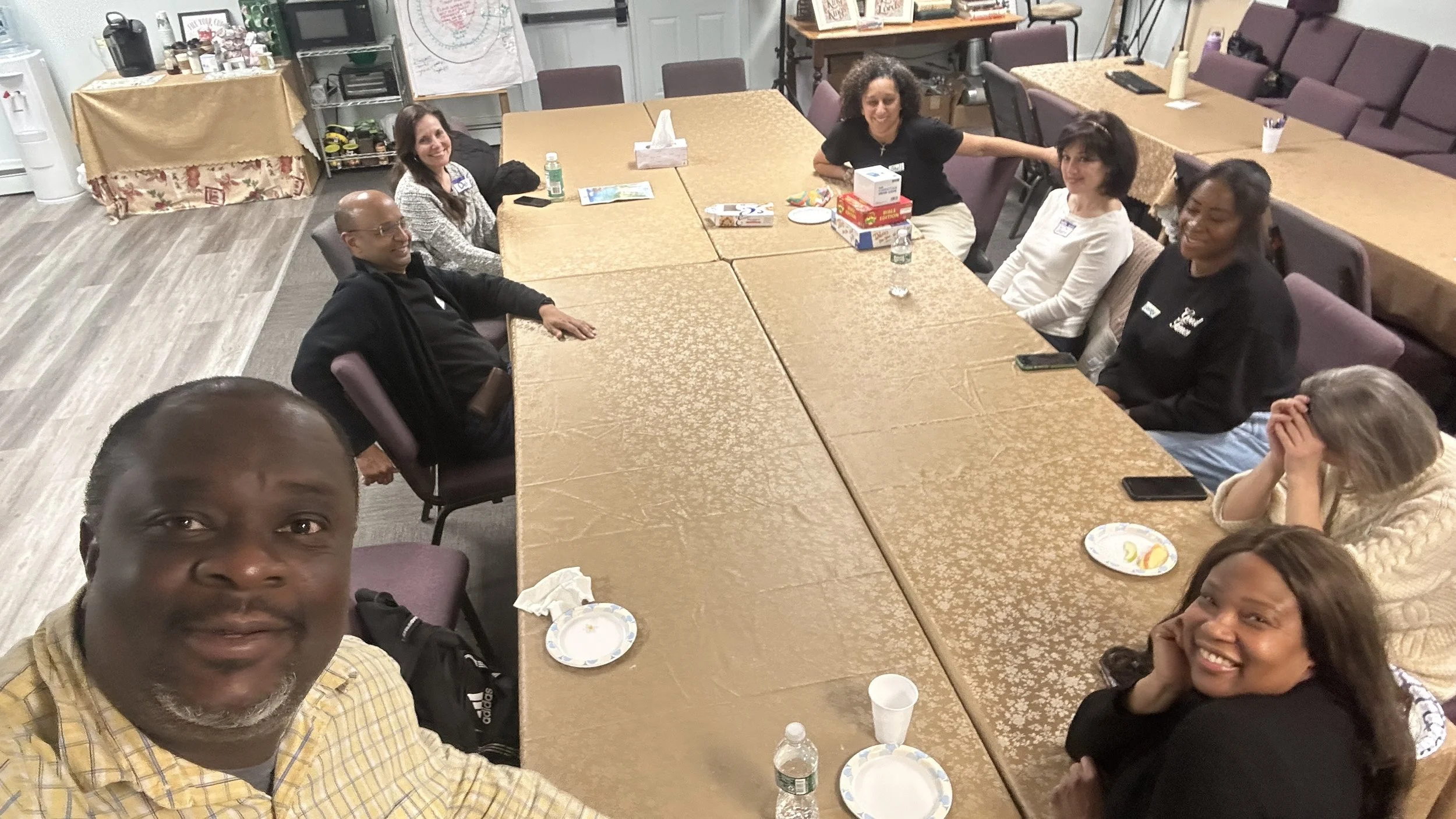 A group of eight diverse people sitting around a large, rectangular table in a meeting or social gathering room, with some plates, bottles of water, and snacks on the table. Two people are taking a selfie at the front of the image, and the others are smiling or covering their face.