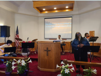 A church service with a woman singing into a microphone, a person playing piano, a woman seated at a table, and a cross on the front of the pulpit, with American flag and flowers in the foreground.