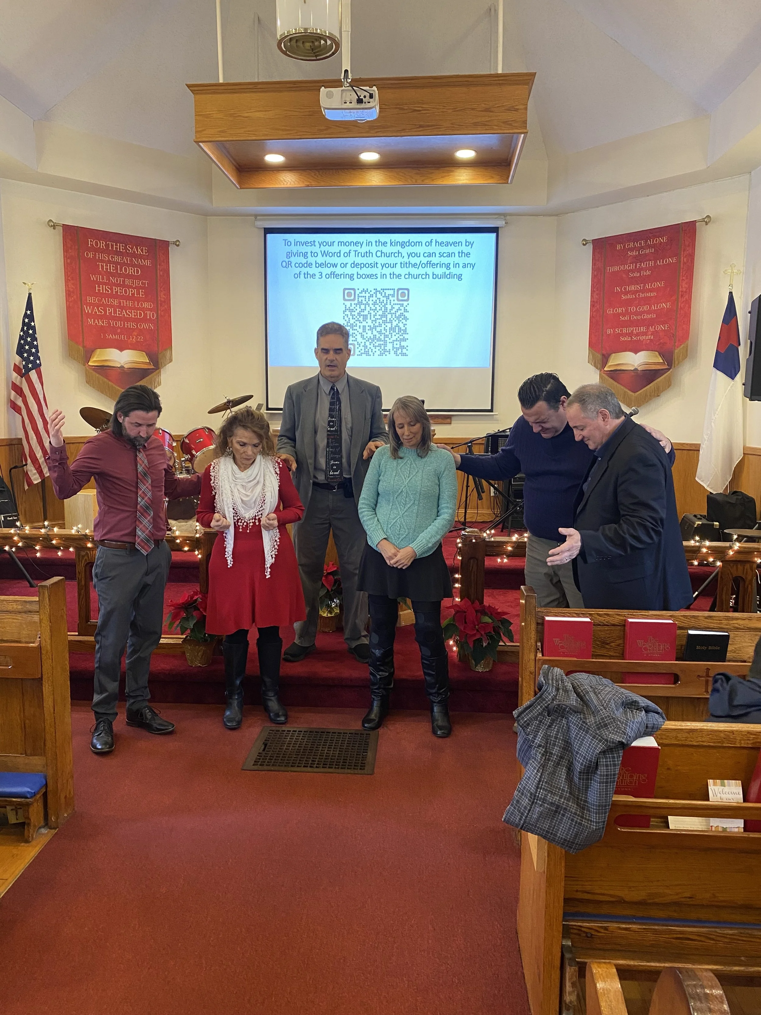 A group of six people praying in a church, with some standing on the altar stage and others in front. The altar is decorated with poinsettias and string lights. A screen behind them displays a QR code for donations.