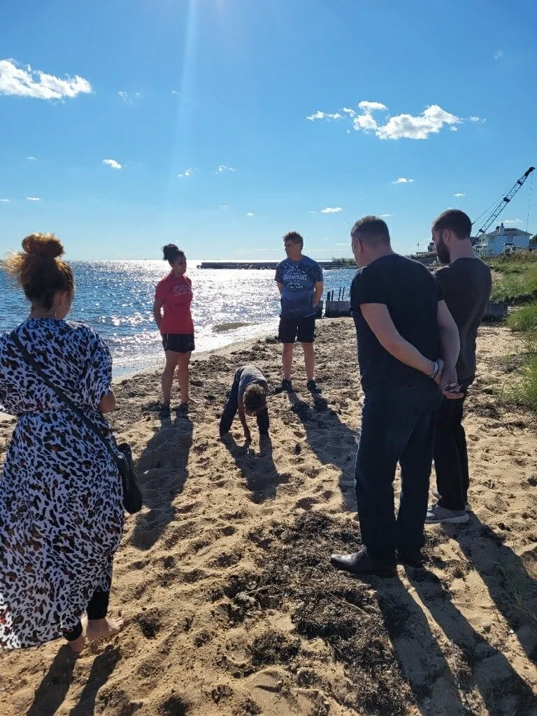 People standing on a sandy beach near the water, with one person bending down and four others observing, under a sunny sky with a few clouds.