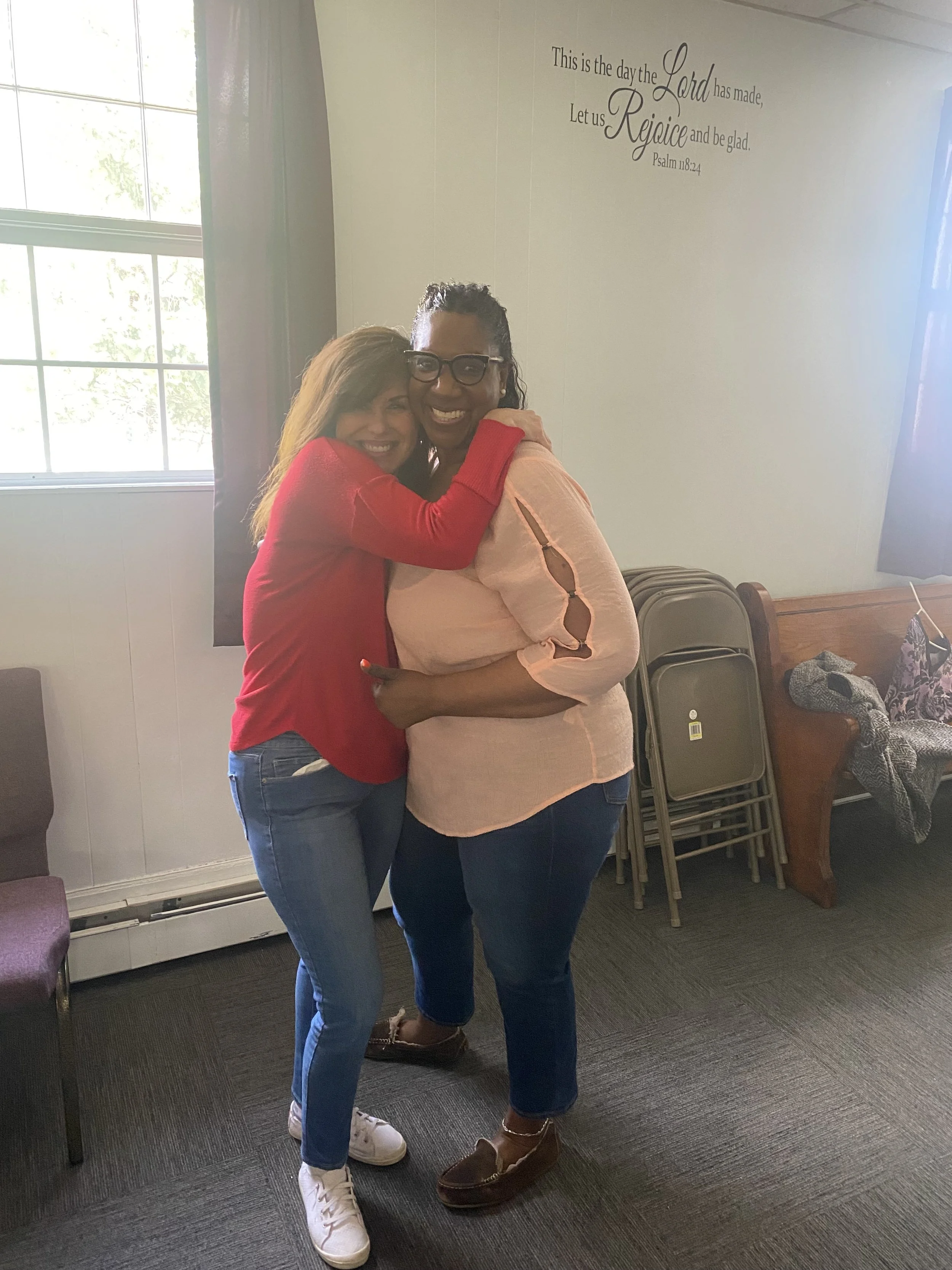 Two women hugging and smiling inside a room, with chairs and a window in the background.