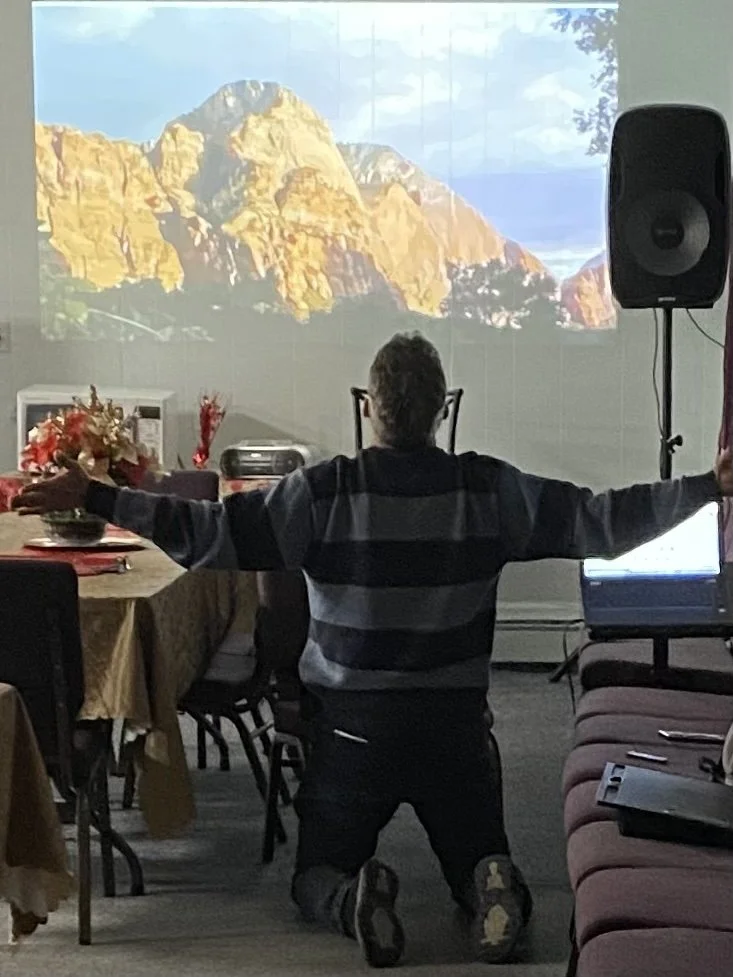 Person kneeling on the floor with arms outstretched in front of a large screen displaying a mountain landscape.