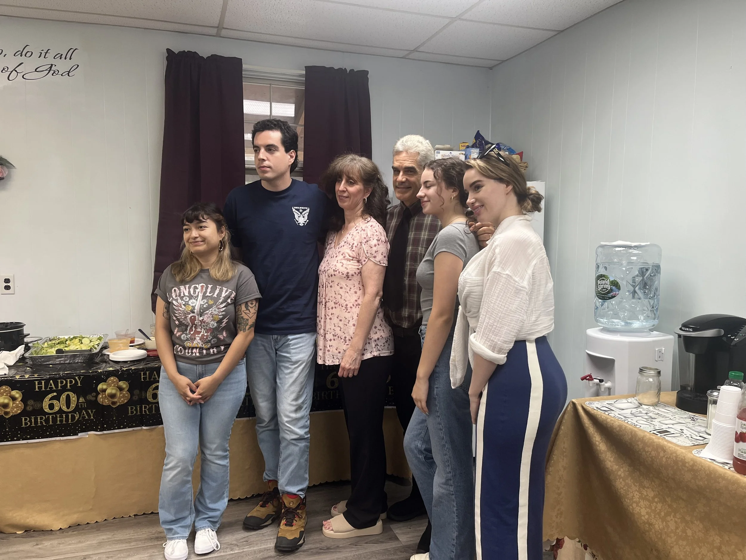 A group of six people, including a young girl, two women, two men, and a young woman, standing together in a room decorated for a birthday celebration. There is a table with food and a 'Happy 60th Birthday' banner in the background, and a water cooler and coffee station on the right.