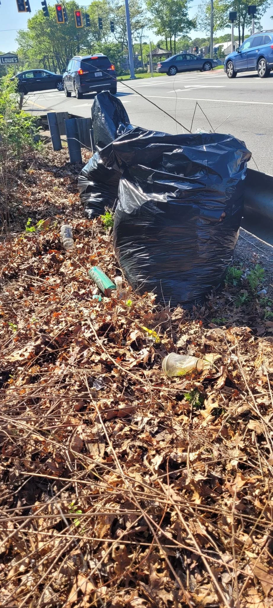 Sidewalk with black trash bags and litter among dry leaves, near a busy street with cars and traffic lights.