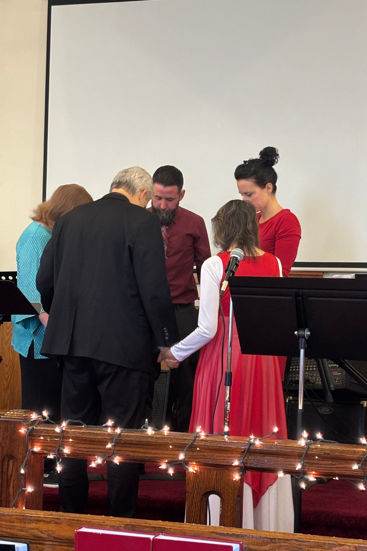 A group of five people standing on a stage holding hands during a ceremony in a church or community hall, with Christmas lights in the foreground and a microphone nearby.