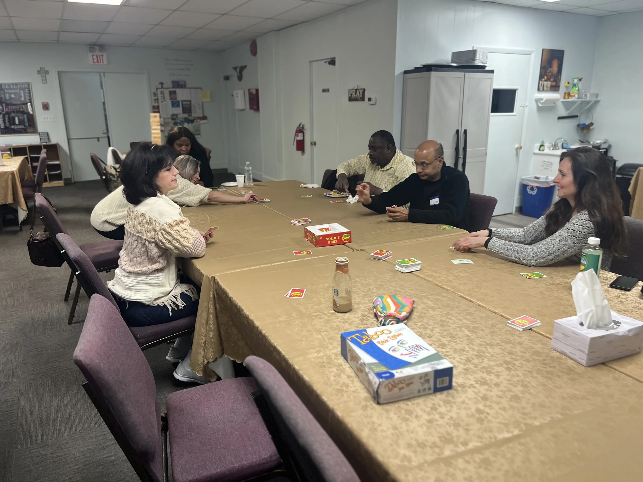 People sitting around a long table playing a card game in a room with religious and community decor.