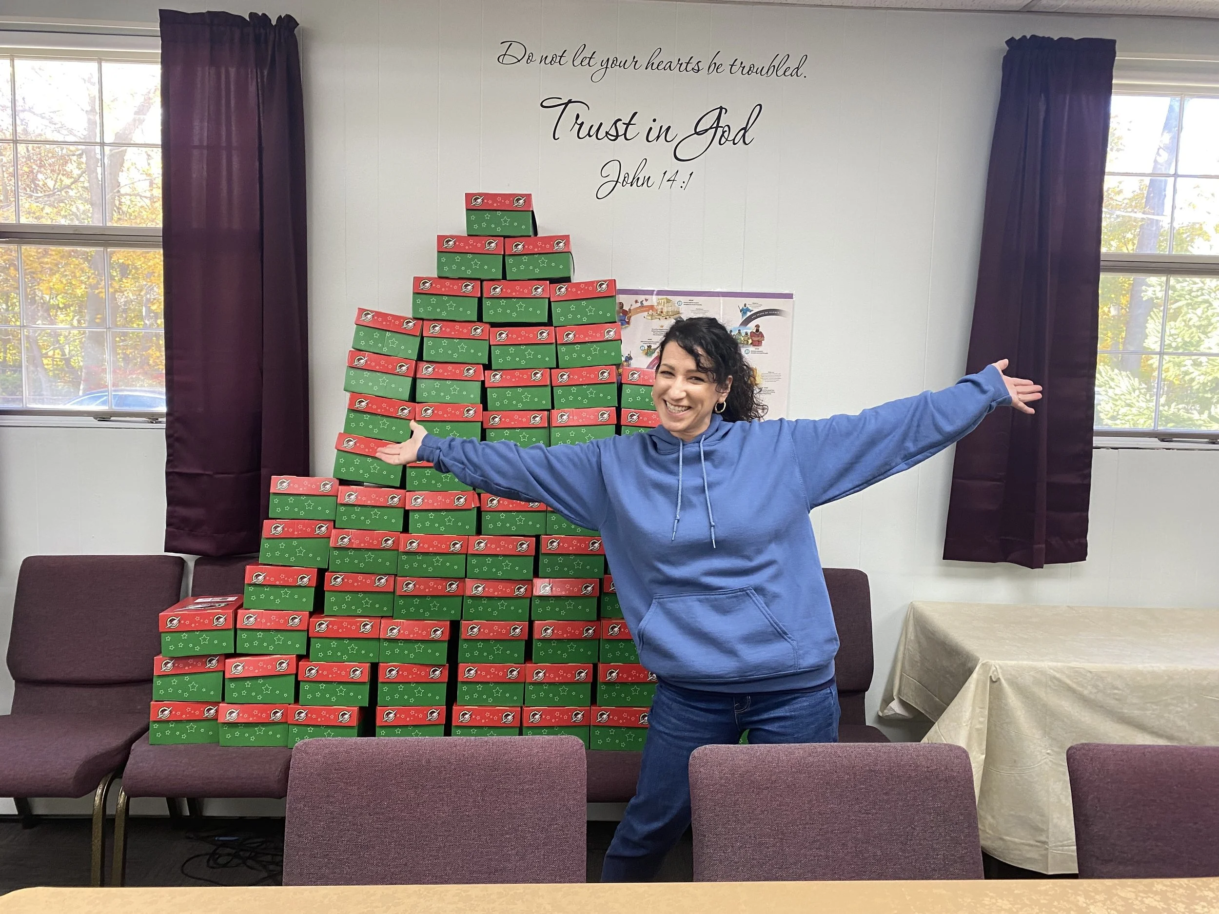 A woman with curly dark hair wearing a blue hoodie and jeans stands in front of a pyramid of stacked Christmas gift boxes and smiles with arms wide open. There are two windows with purple curtains, a wall with a Bible quote, and chairs and a table in the room.