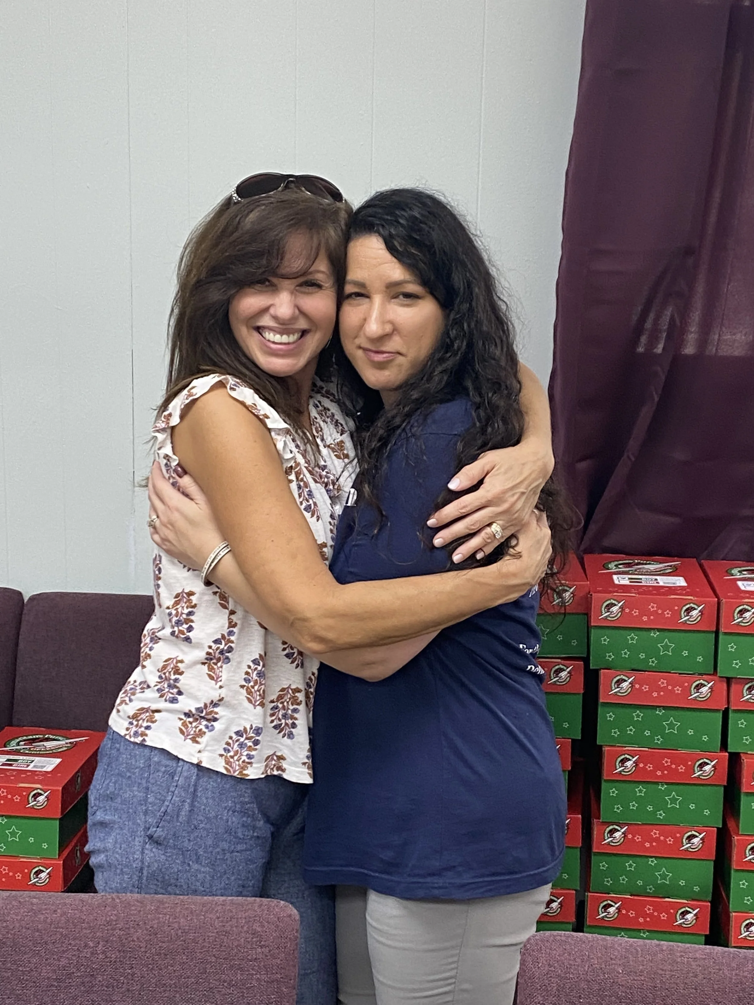 Two women hugging and smiling in front of stacked pizza boxes, one wearing a sleeveless floral blouse and the other in a navy blue shirt.