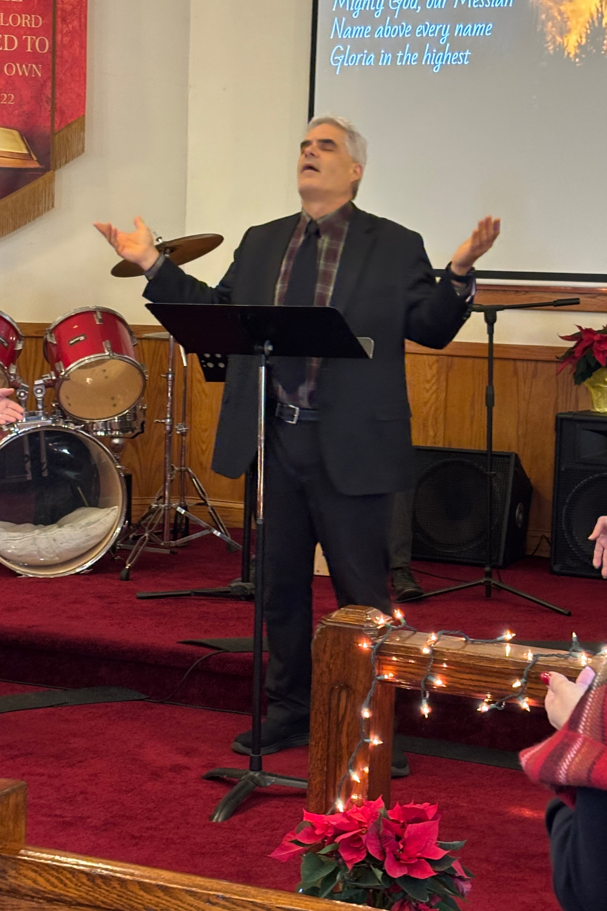 A man with gray hair and a black suit is standing on a stage, raising his hands in a prayer or worship gesture, with his eyes closed. There are drums behind him and a screen displaying lyrics above. The setting appears to be a church or worship service, decorated with poinsettias and Christmas lights.