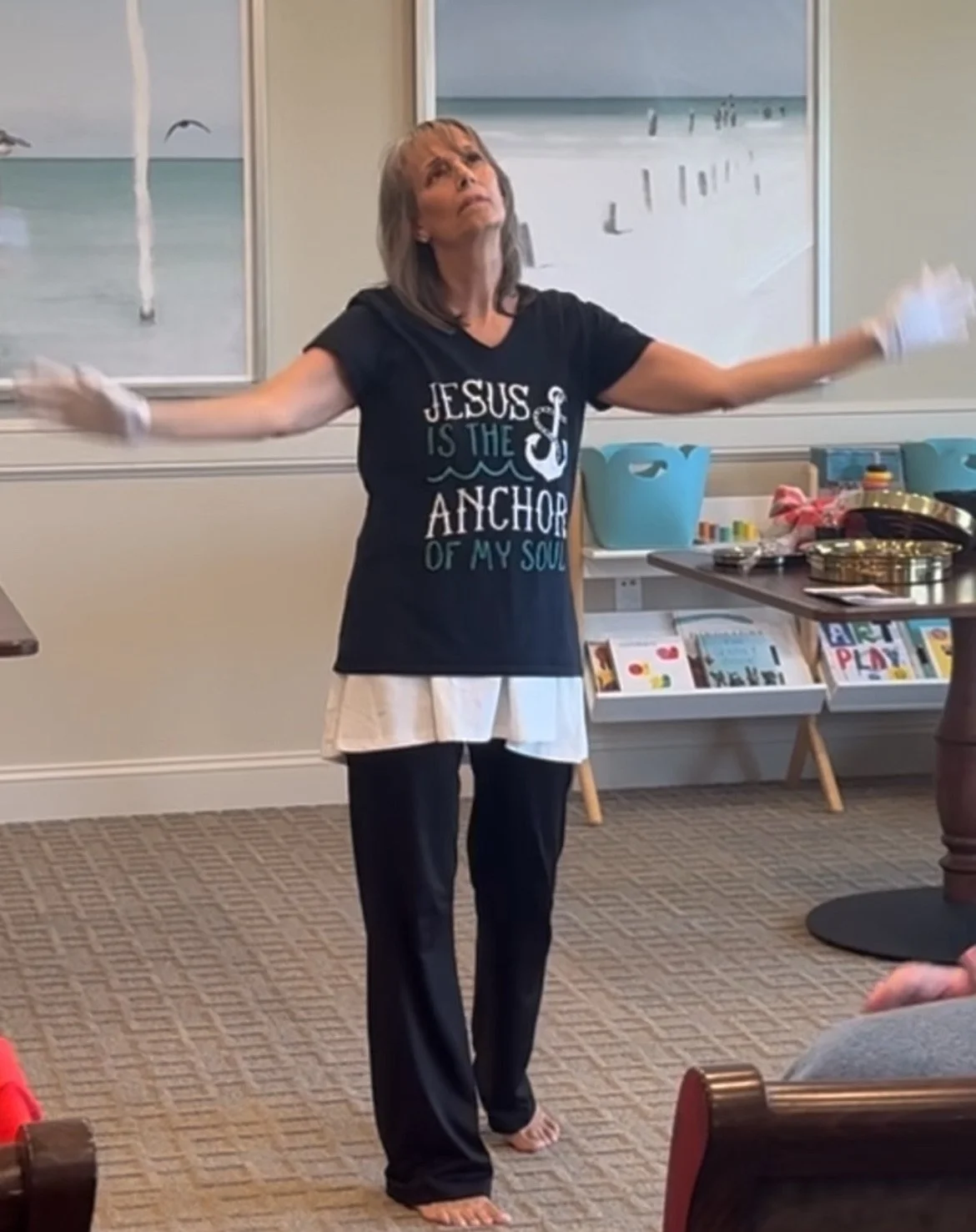 A woman with gray hair stands with her arms outstretched, wearing a black T-shirt that reads "Jesus is the anchor of my soul" and white gloves indoors, with a beach-themed painting in the background.