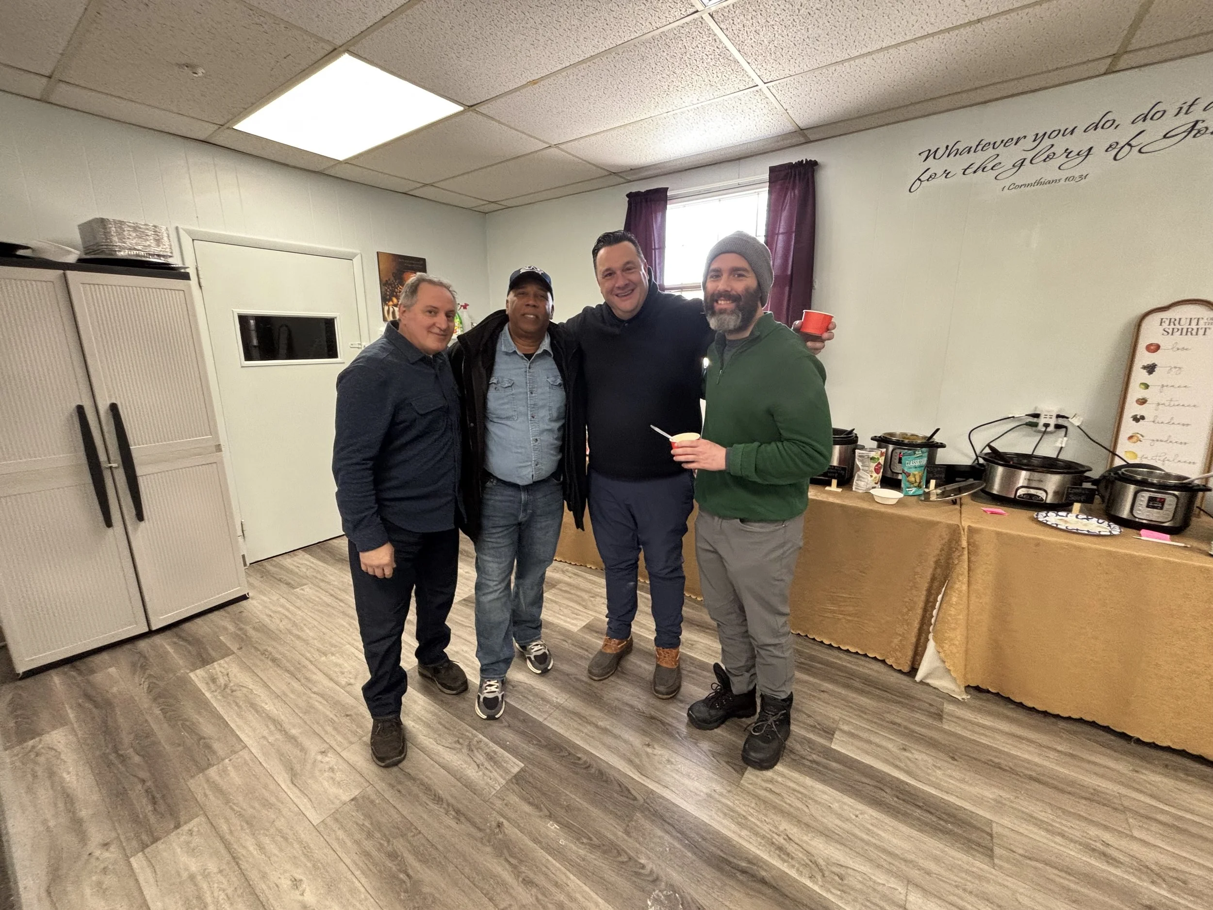 Four men standing together in a room, smiling and holding cups, with a buffet table in the background.