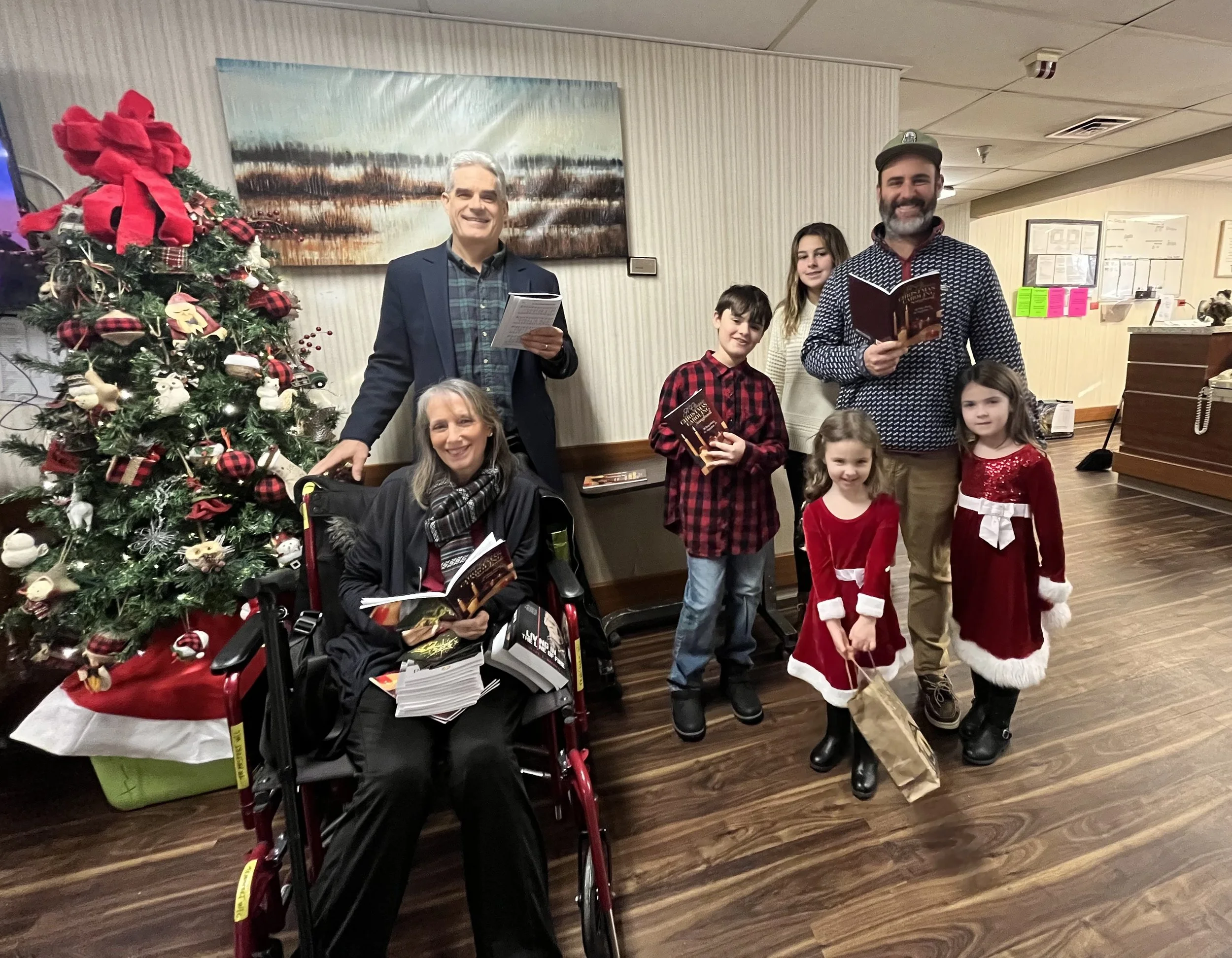 Group of people celebrating Christmas indoors with a decorated Christmas tree. Two adults, three children, and a woman in a wheelchair are smiling and holding books or gift bags.