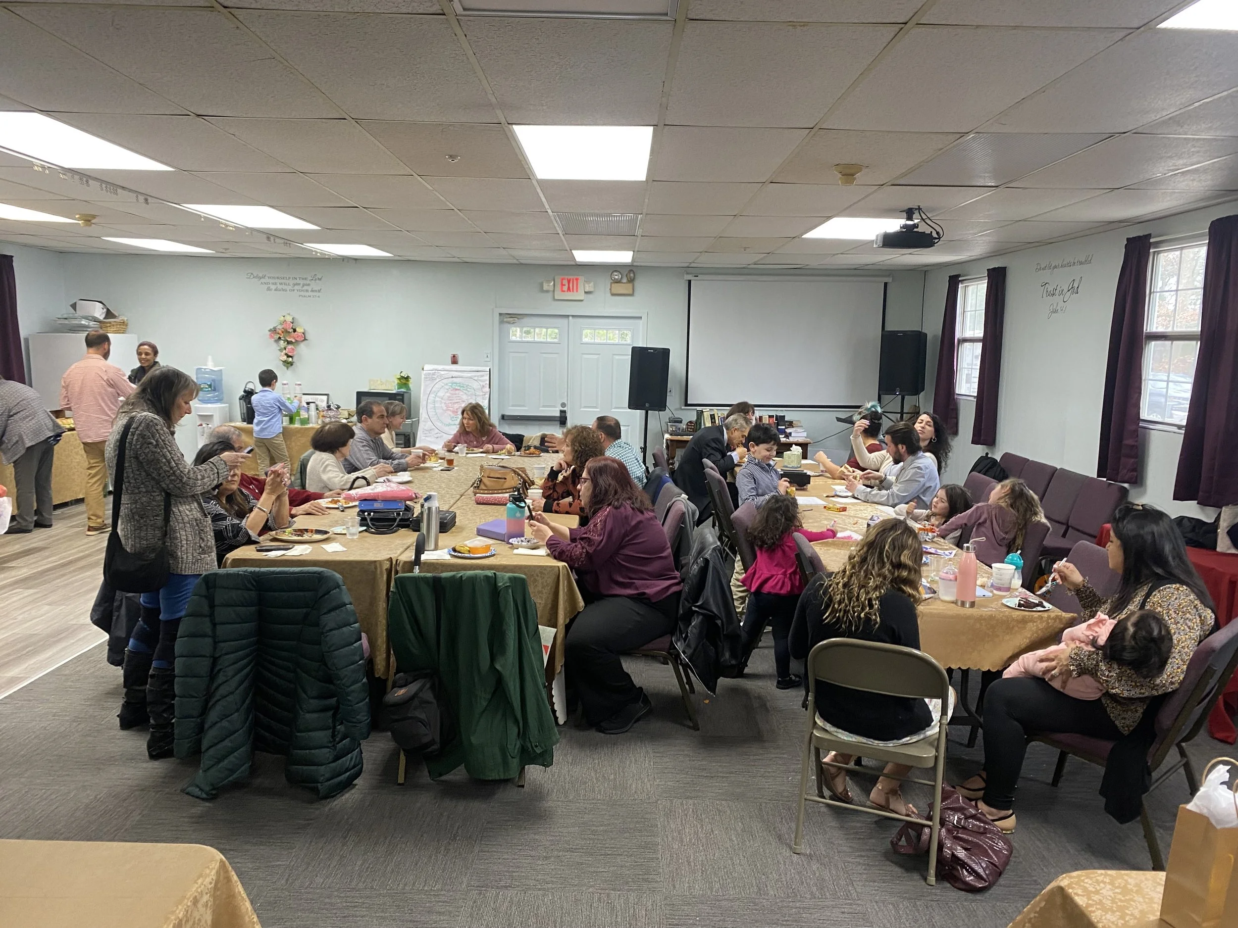 People sitting around tables in a room, participating in a gathering or event, with some standing and chatting, and a water dispenser and projector in the background.