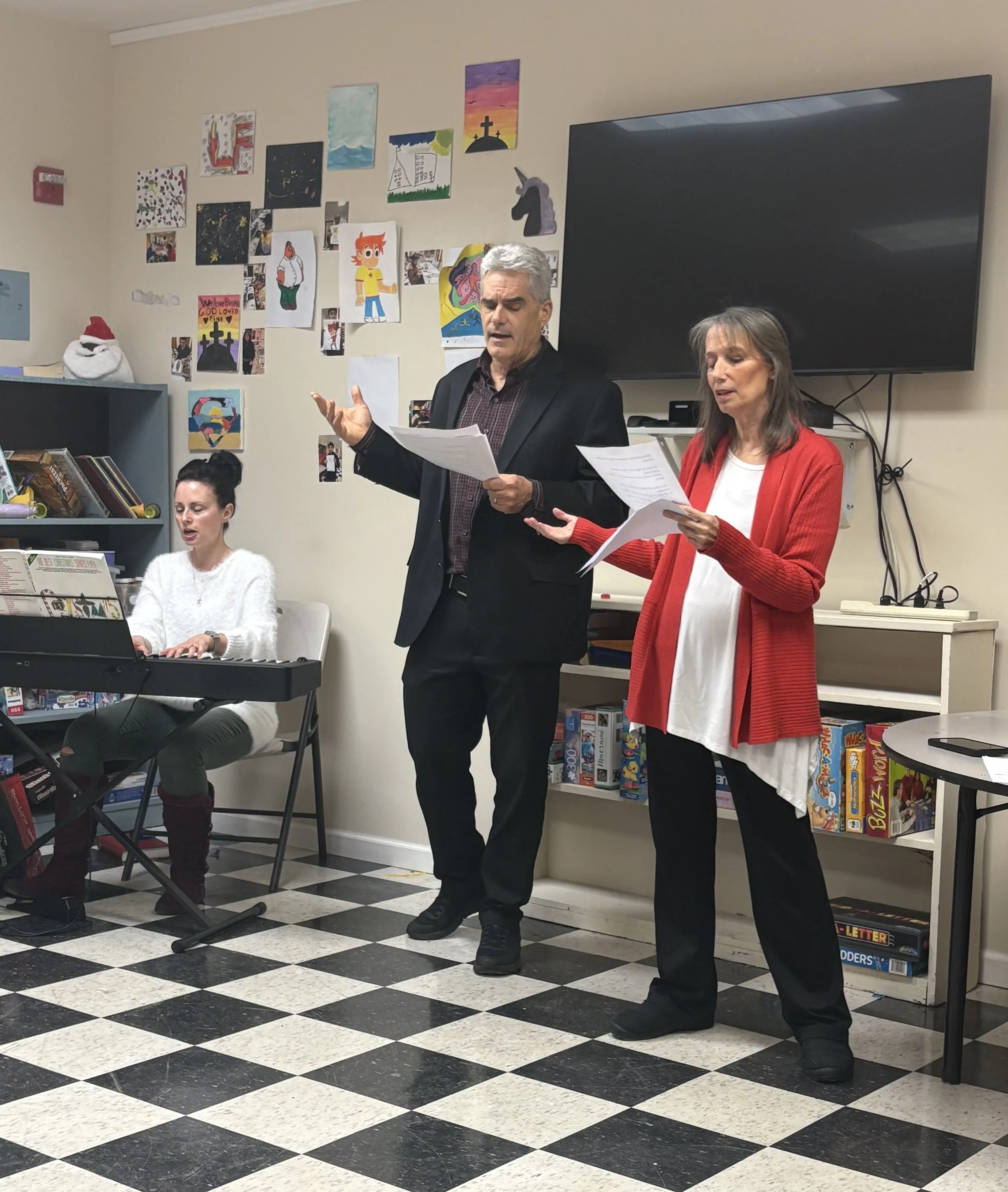 Three people singing or performing in a room decorated with children's artwork. One woman is playing the keyboard, while the man and woman stand with papers in hand, singing or speaking. The room has black and white checkered flooring and shelves with board games.