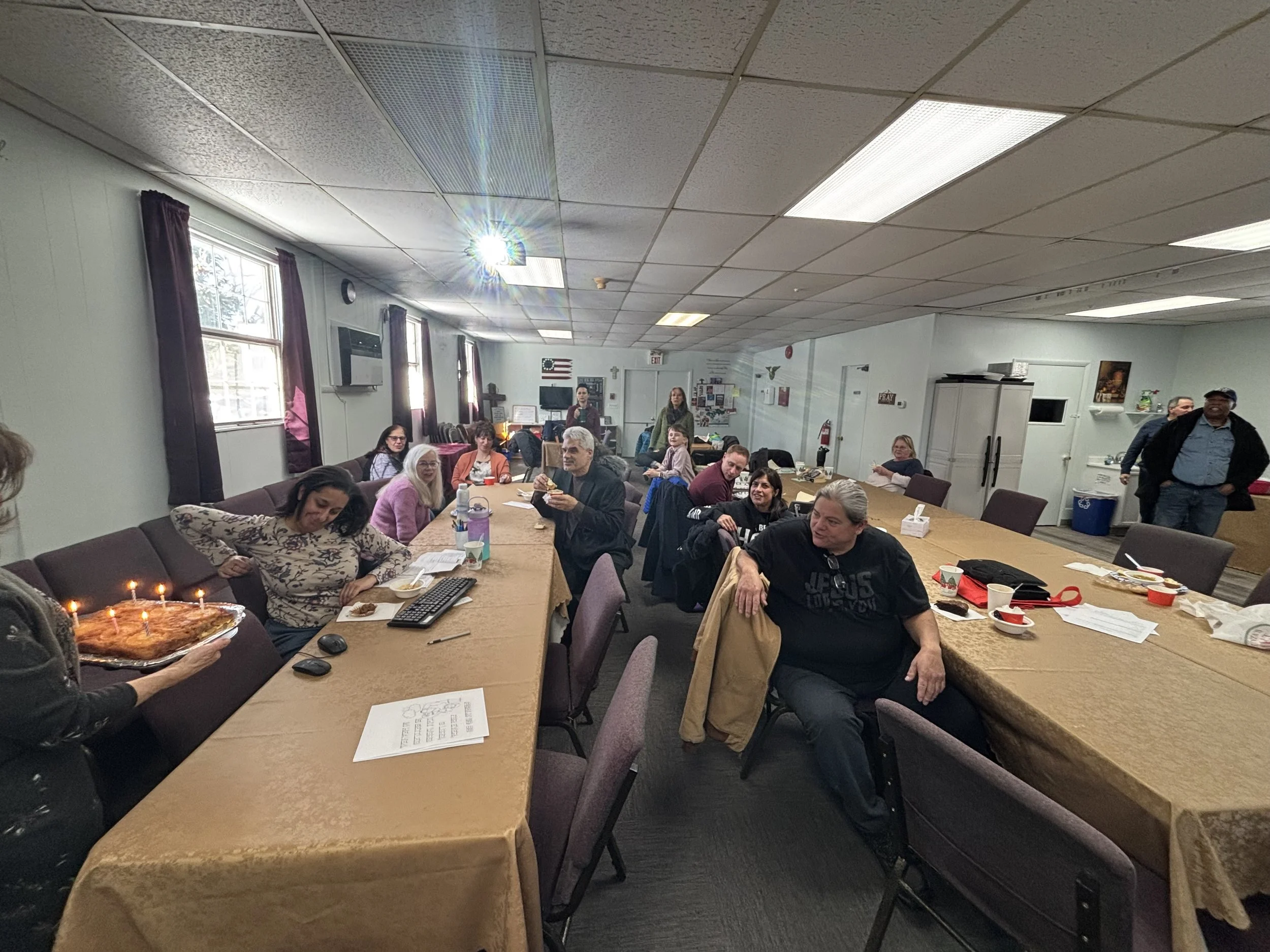 Group of people gathered in a room celebrating a birthday with a cake that has lit candles. Some people are seated at tables, while others stand and chat.