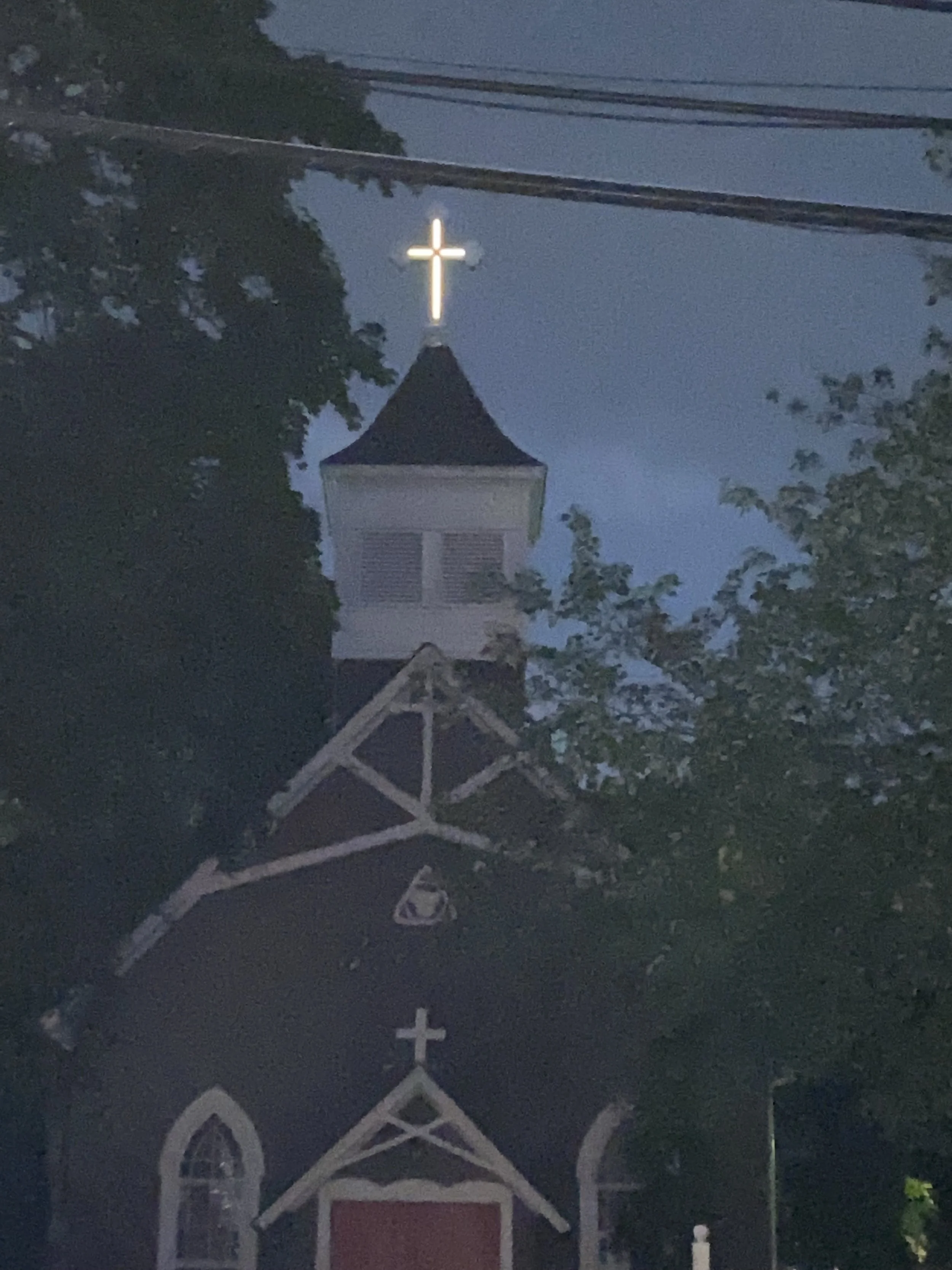 Nighttime view of a church with a steeple topped by a lit cross, surrounded by trees and power lines.