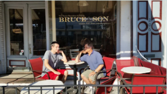 Two young men sitting outside a cafe, talking at a small table with red and white chairs.