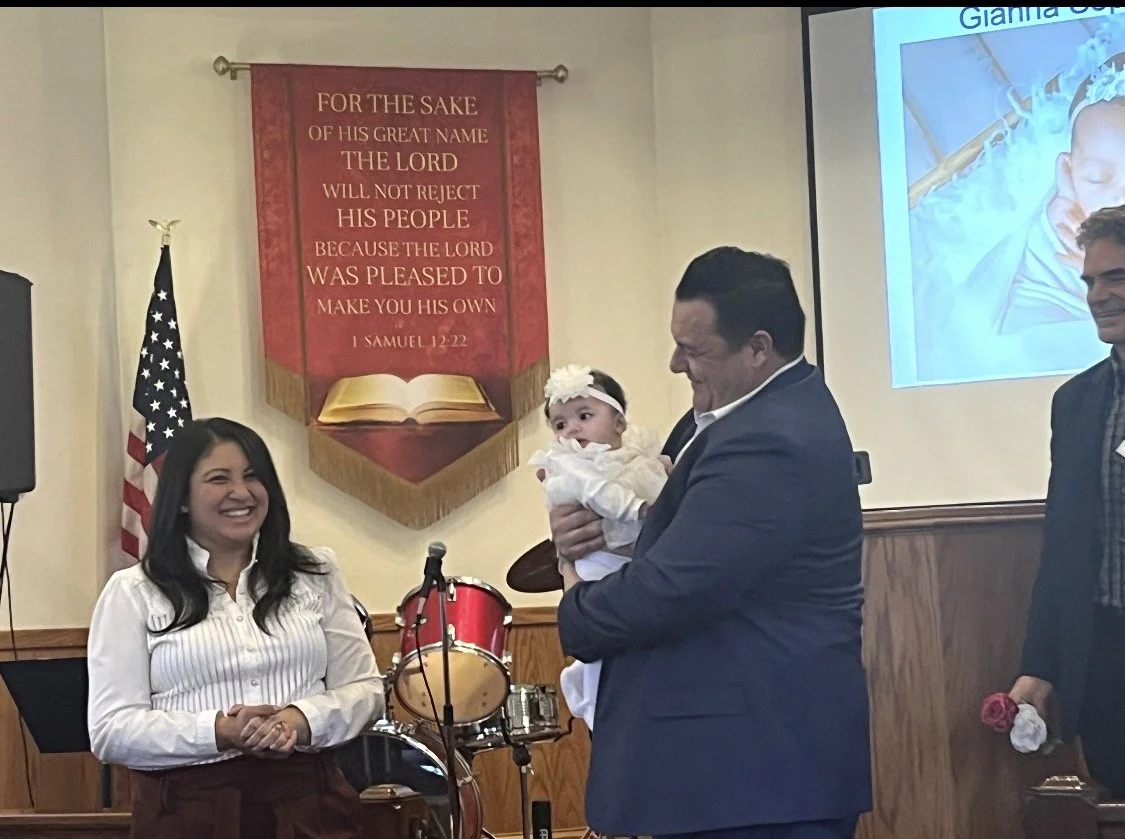 A man in a blue suit holding a baby girl dressed in white during a religious ceremony, with a woman smiling nearby. Behind them, a red banner with gold lettering and an American flag are displayed, along with a drum set and a projection of an image of Jesus Christ on the wall.