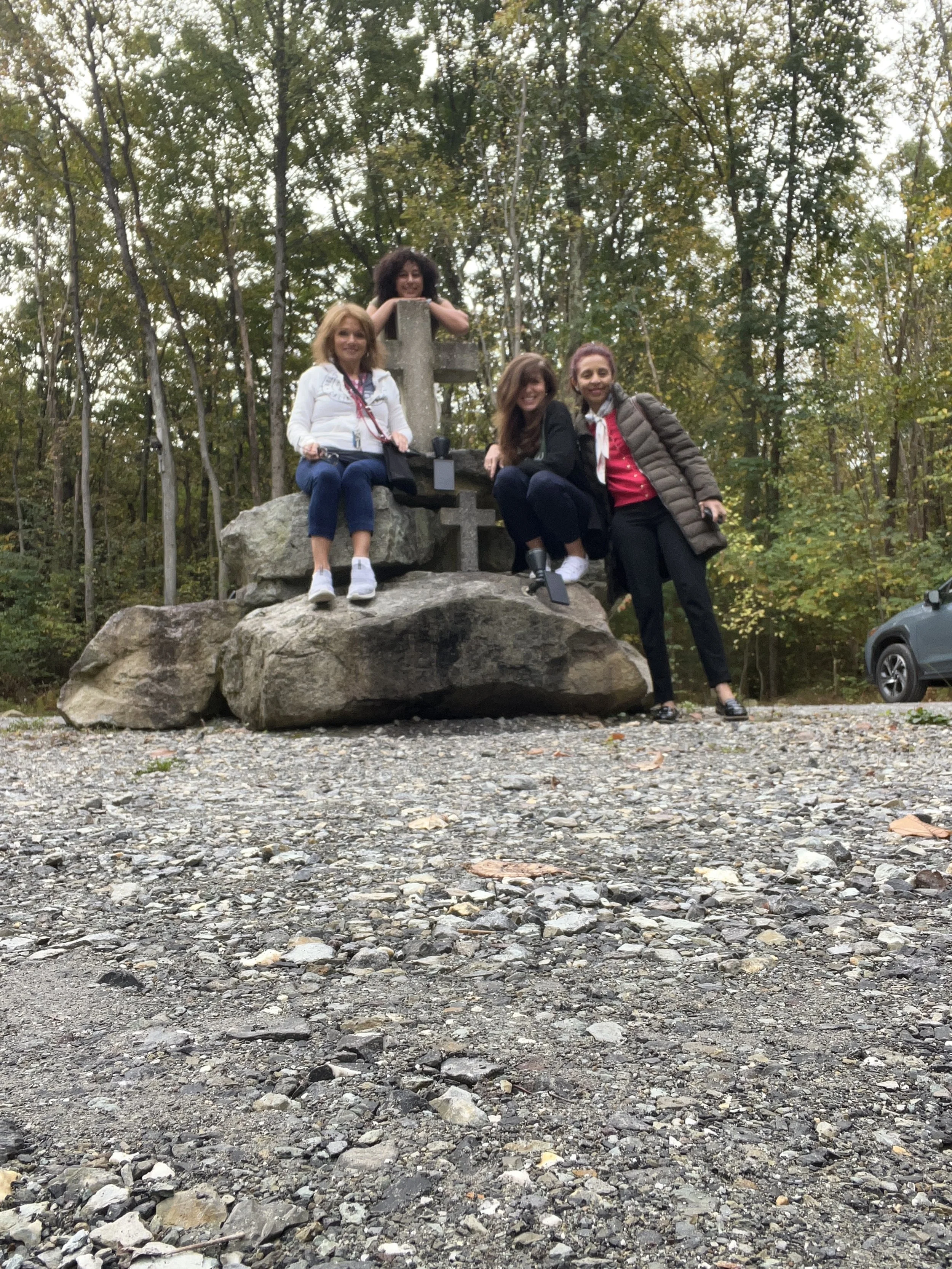 Four women sitting and standing on a large rock with a wooden cross, surrounded by trees in a forested area.
