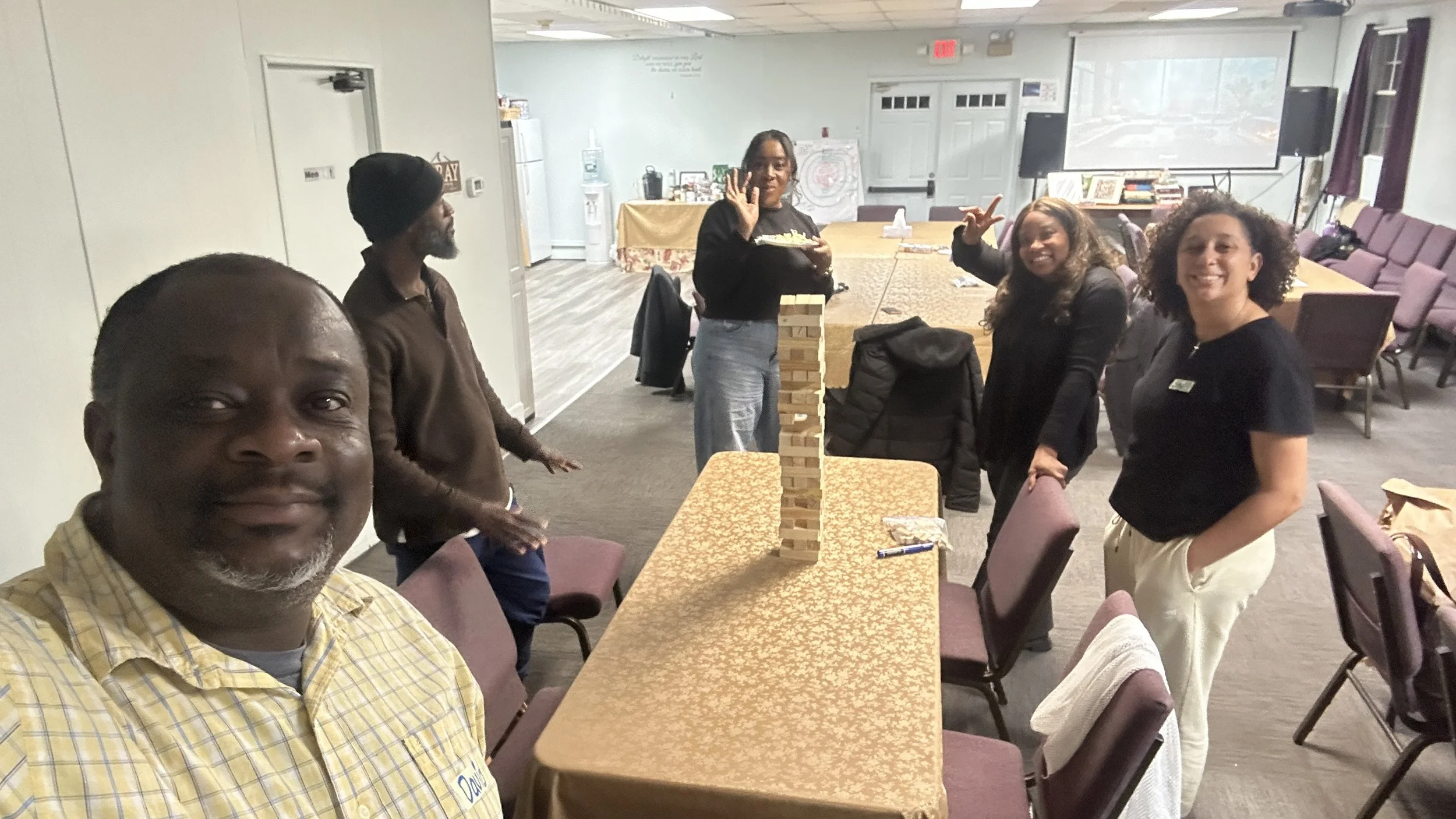 Group of people playing giant Jenga game in a meeting room with a long table and chairs, some of them smiling and enjoying the game.