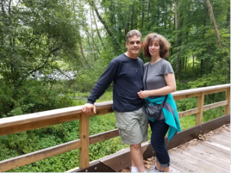 A man and woman standing on a wooden bridge in a forested area, smiling at the camera.