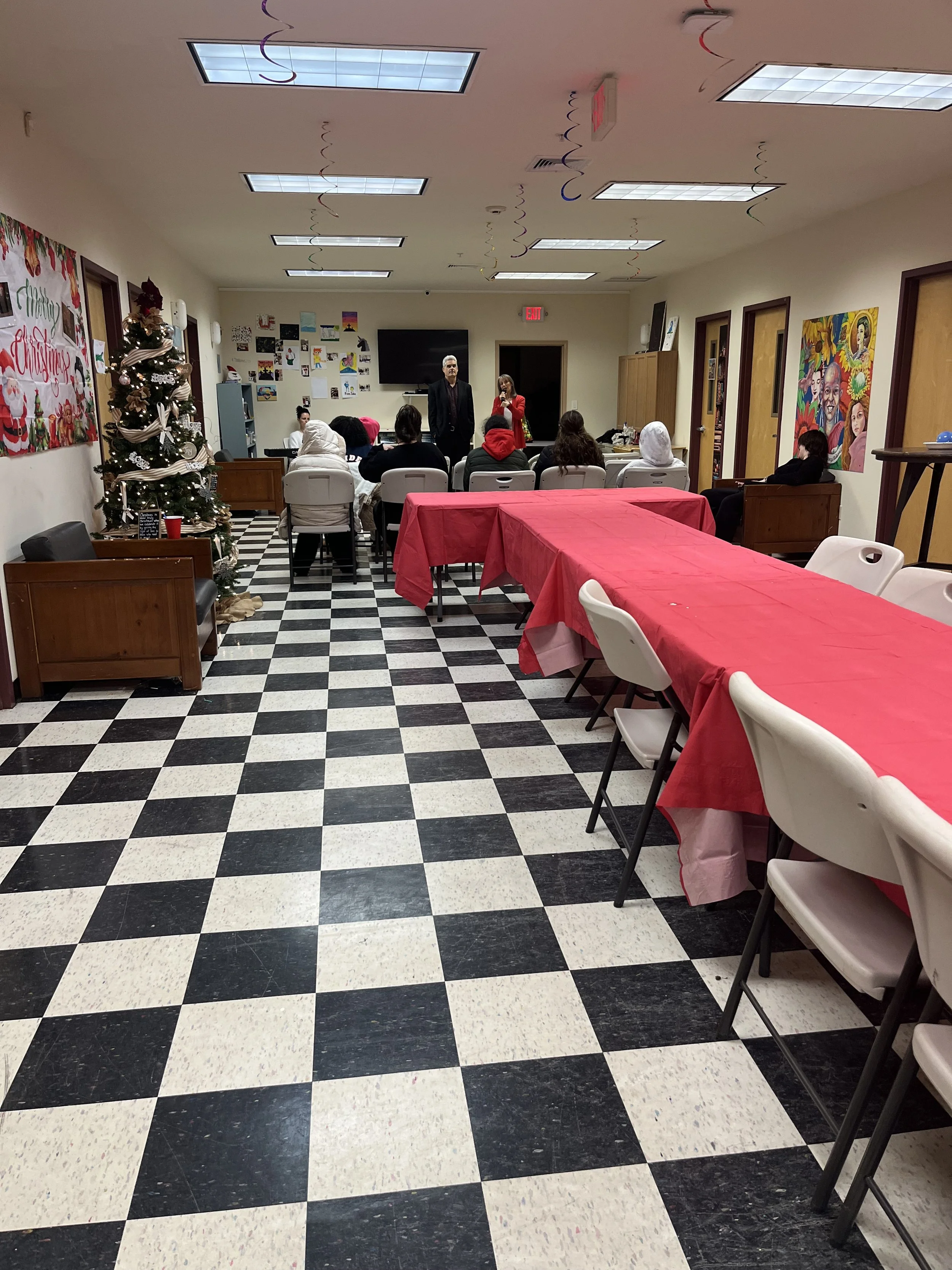 A decorated room for a Christmas event with a Christmas tree, red tablecloths, and people seated at tables, some wearing winter clothing, facing a small stage area with two people speaking.