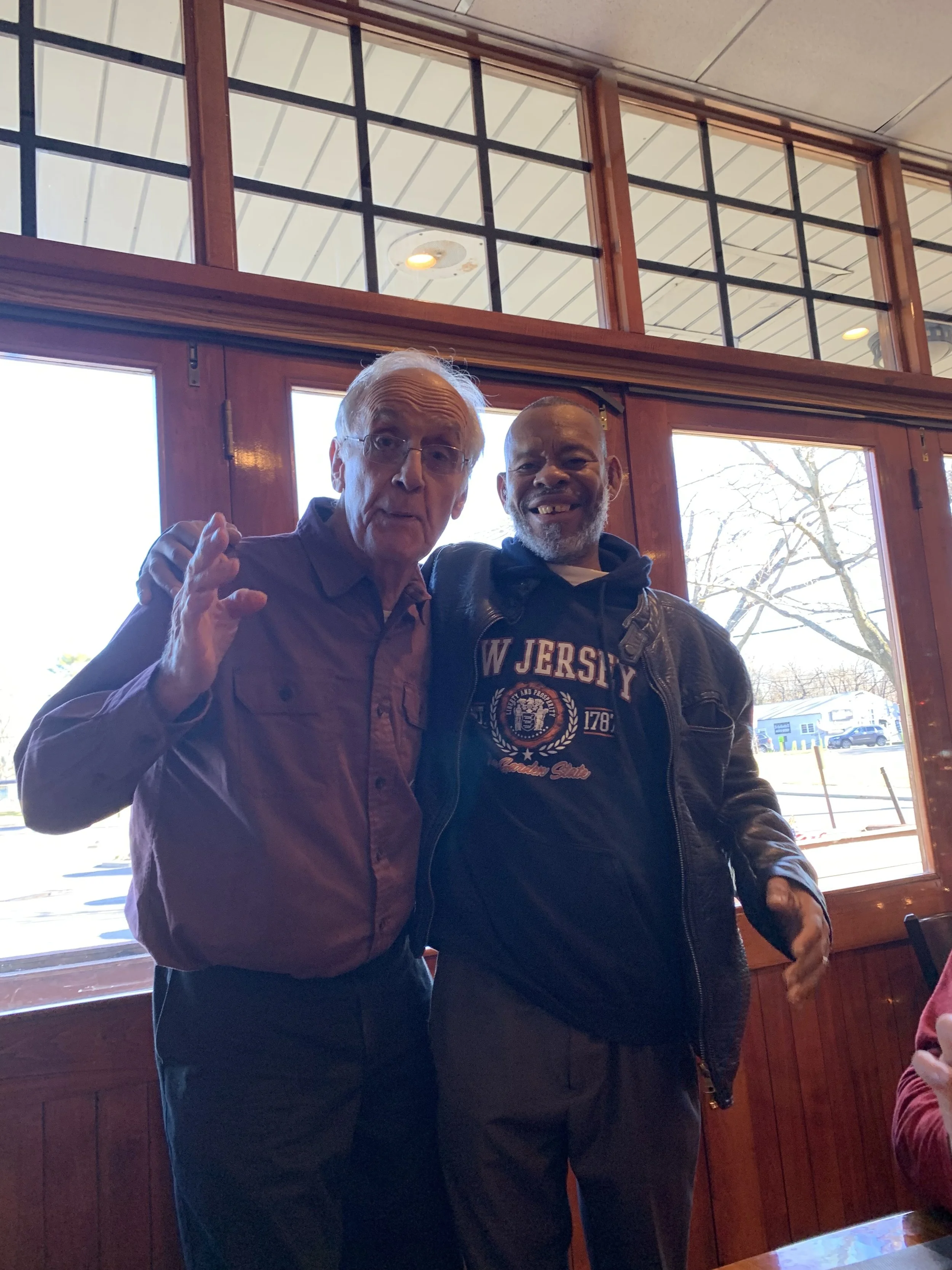 Two men standing inside a restaurant near large windows. The man on the left is older with light skin, gray hair, glasses, and wearing a purple button-up shirt. The man on the right is younger with dark skin, short hair, a beard, and wearing a black hoodie with 'W Jersey' printed on it and a black leather jacket. They are smiling and posing for the photo.