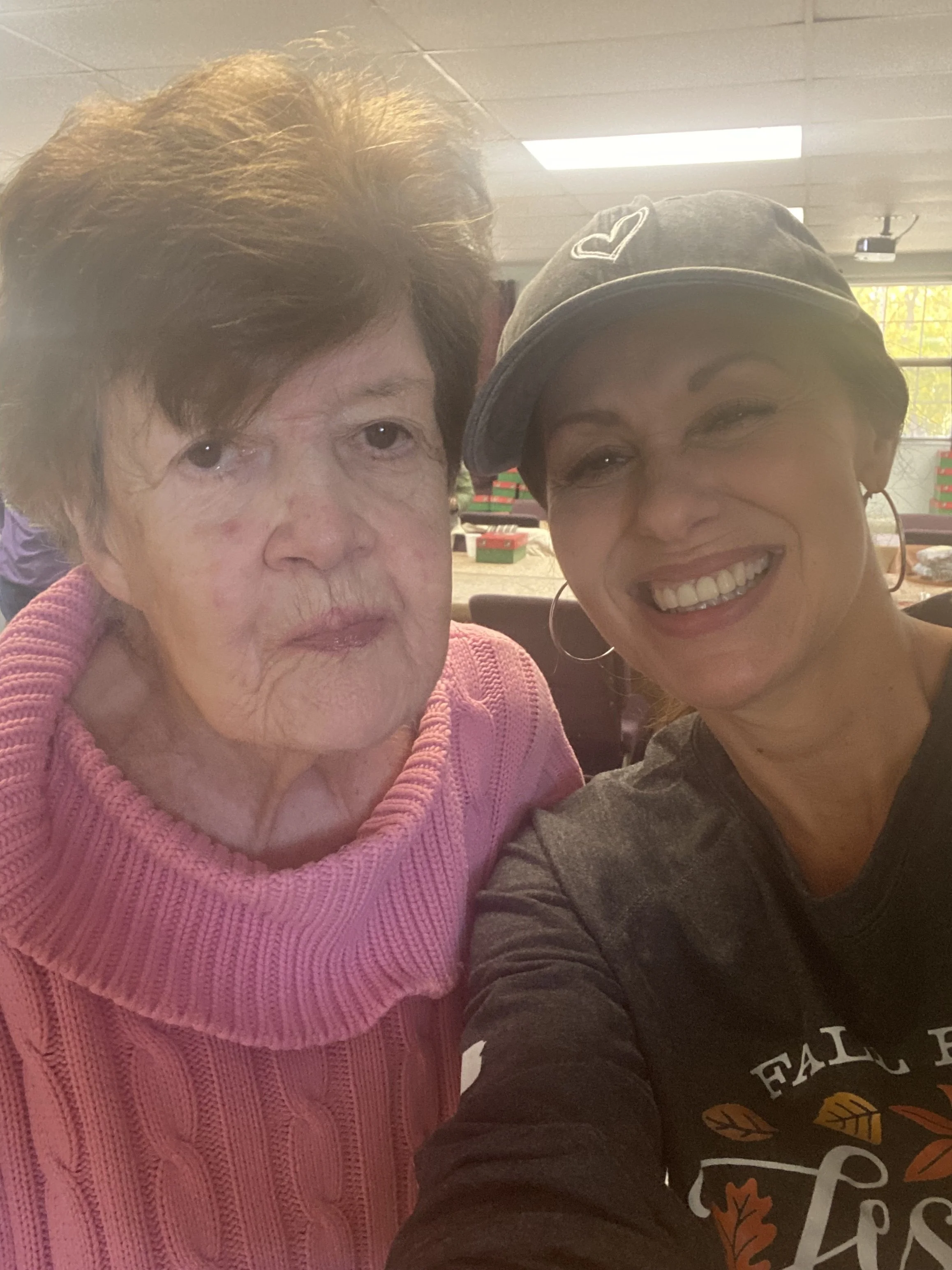 Two women taking a close-up selfie indoors, with one older woman wearing a pink sweater and the other woman wearing a dark shirt, hoop earrings, and a cap, both smiling and looking at the camera.