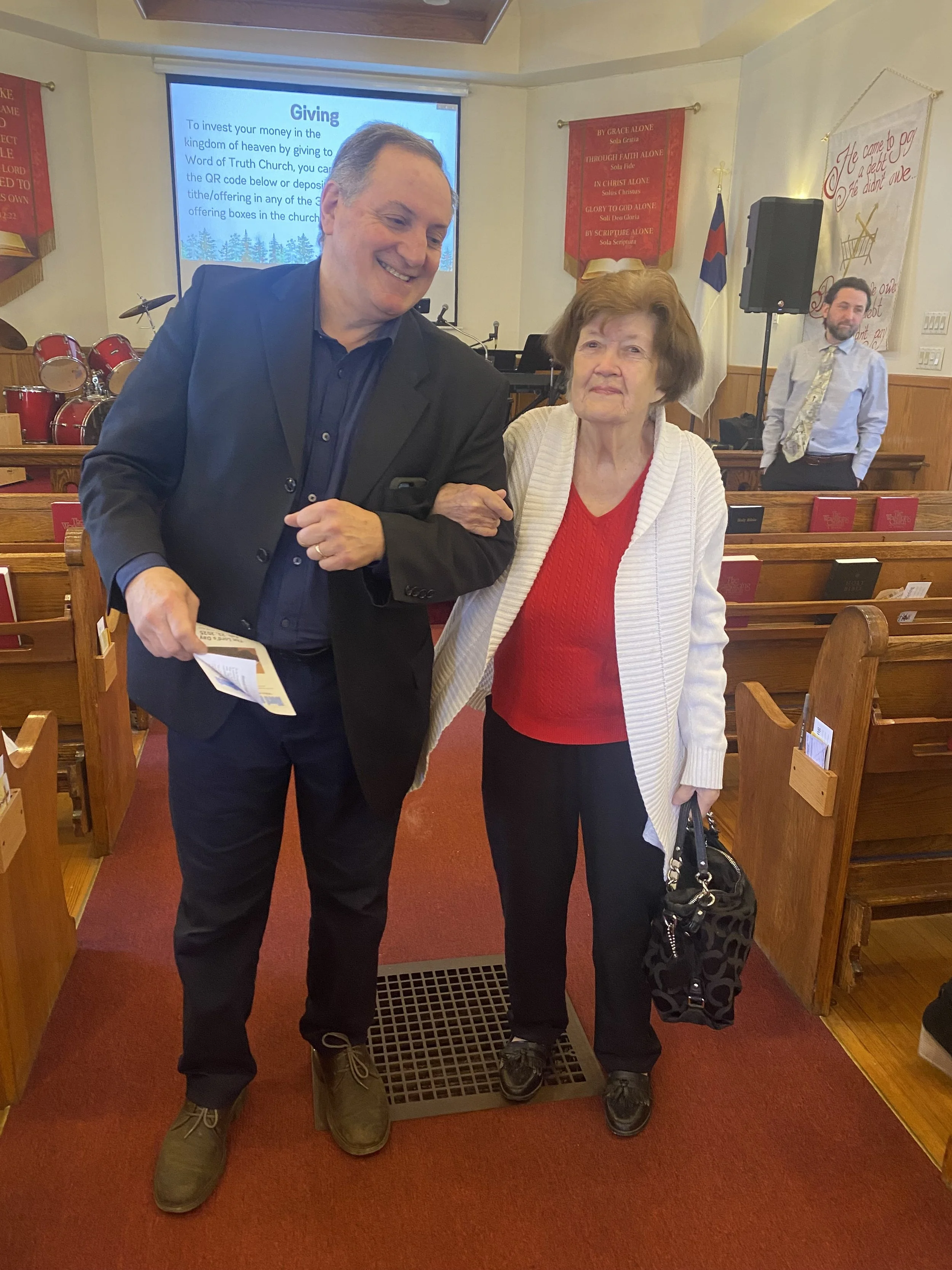 A man and an elderly woman are standing together inside a church, with a woman in the background and a large screen displaying a religious message behind them.