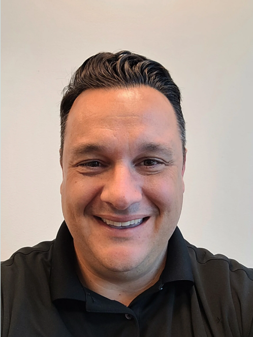 A smiling man with dark hair styled back, wearing a black collared shirt, standing in front of a plain light-colored wall.