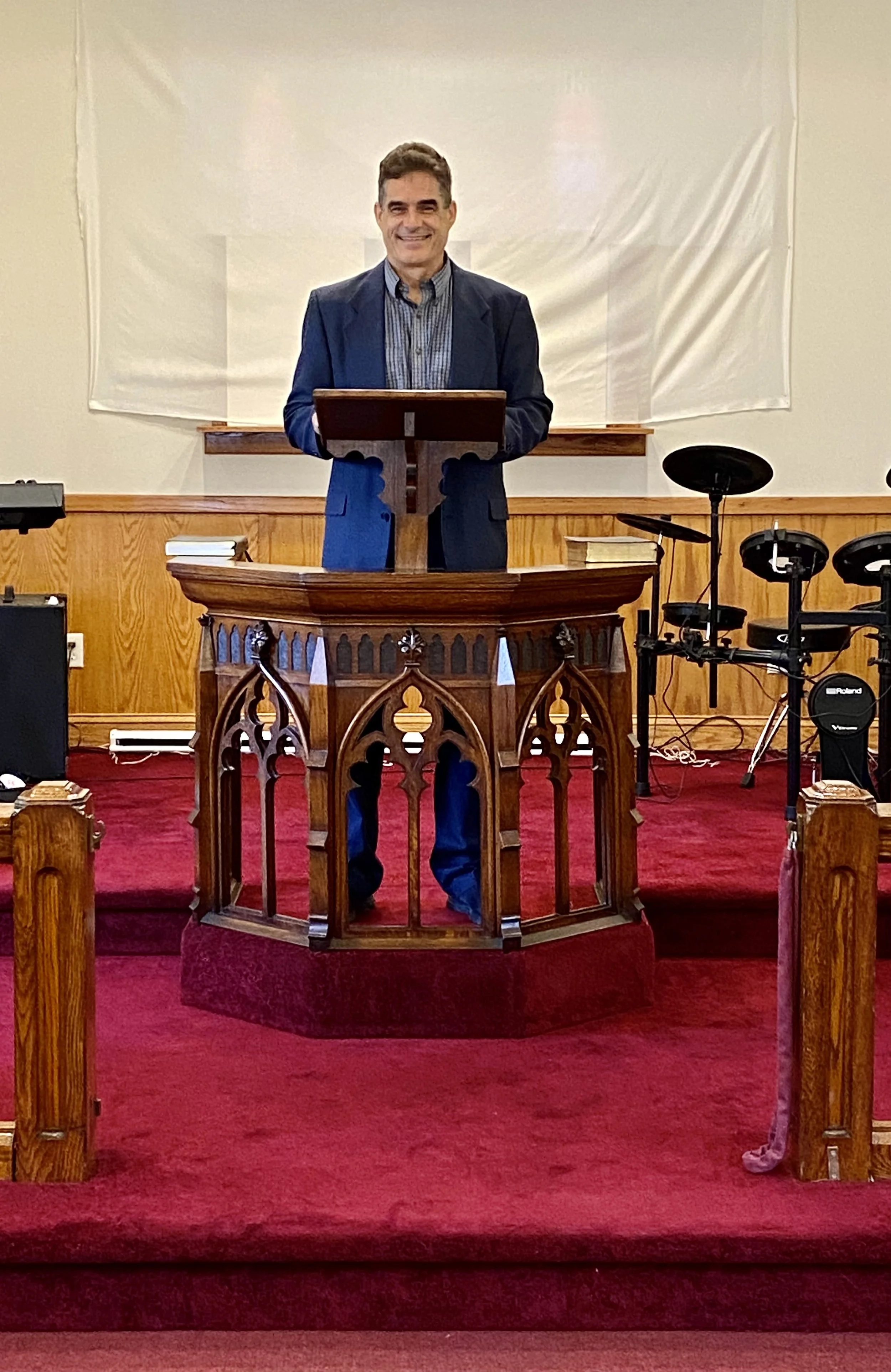 A man standing at a wooden pulpit in a church, smiling, with musical instruments on the right side and a white banner in the background.