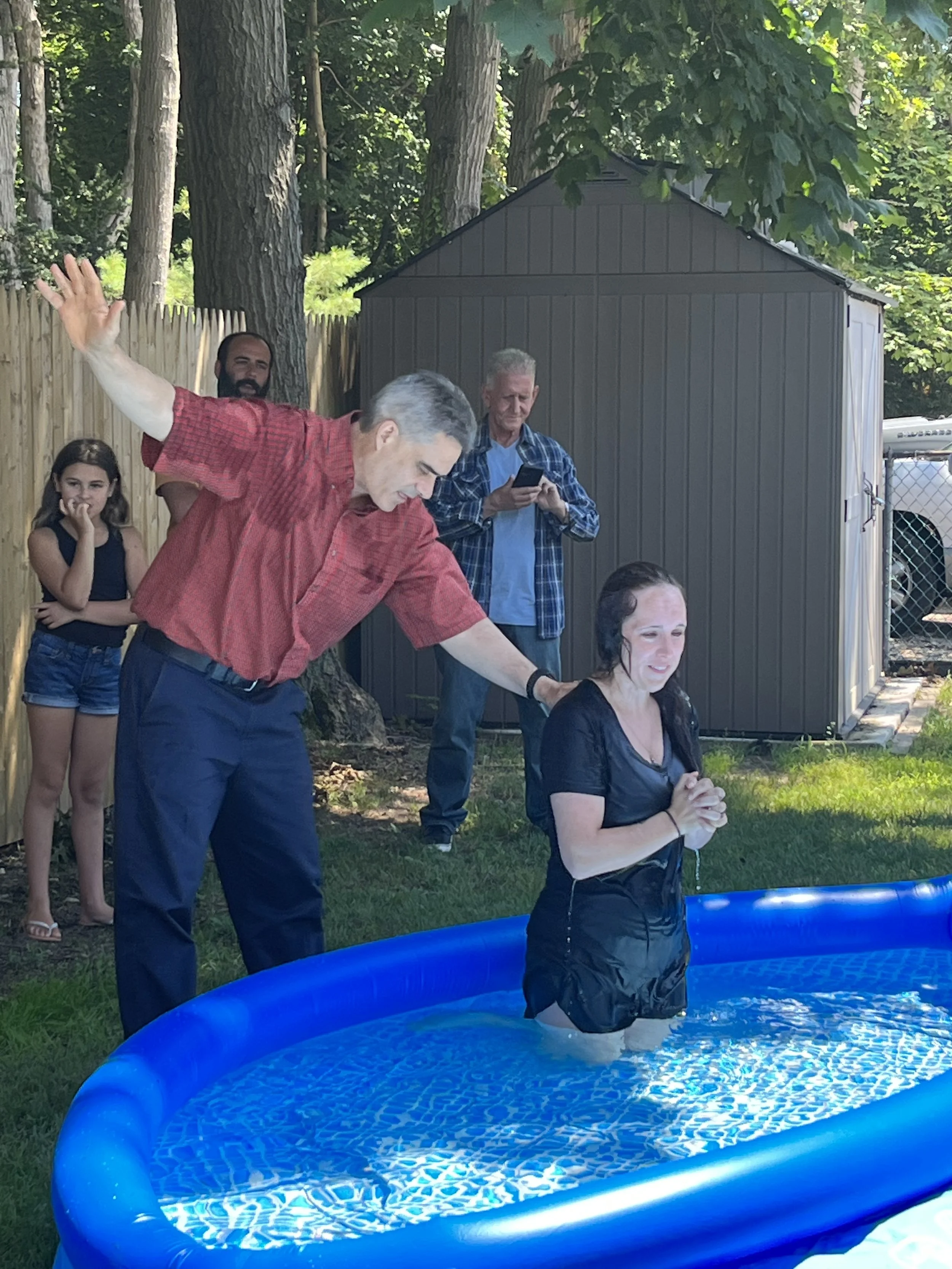 A woman standing in an inflatable pool, praying, while a man is pouring water on her head during an outdoor religious baptism ceremony with four onlookers in the background.