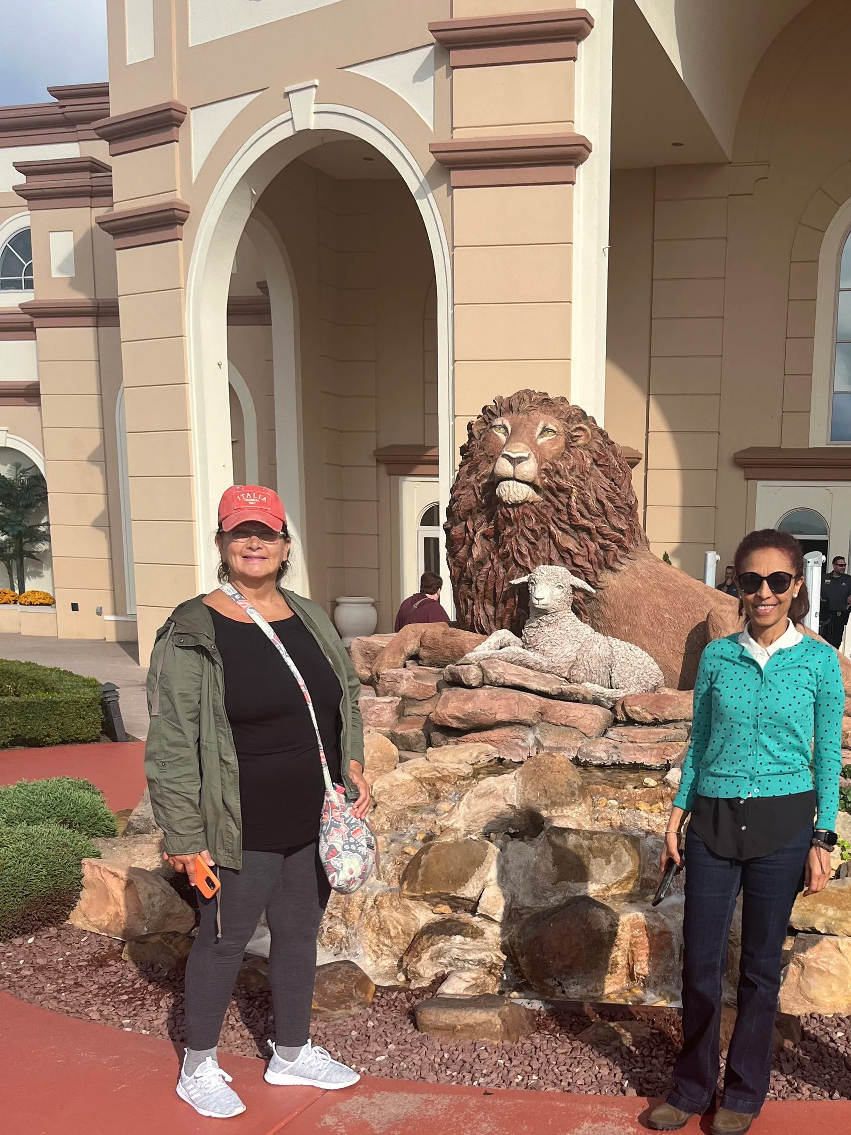 Two women standing in front of a large lion statue with a smaller sheep statue on rocks, in front of a grand building with arches.