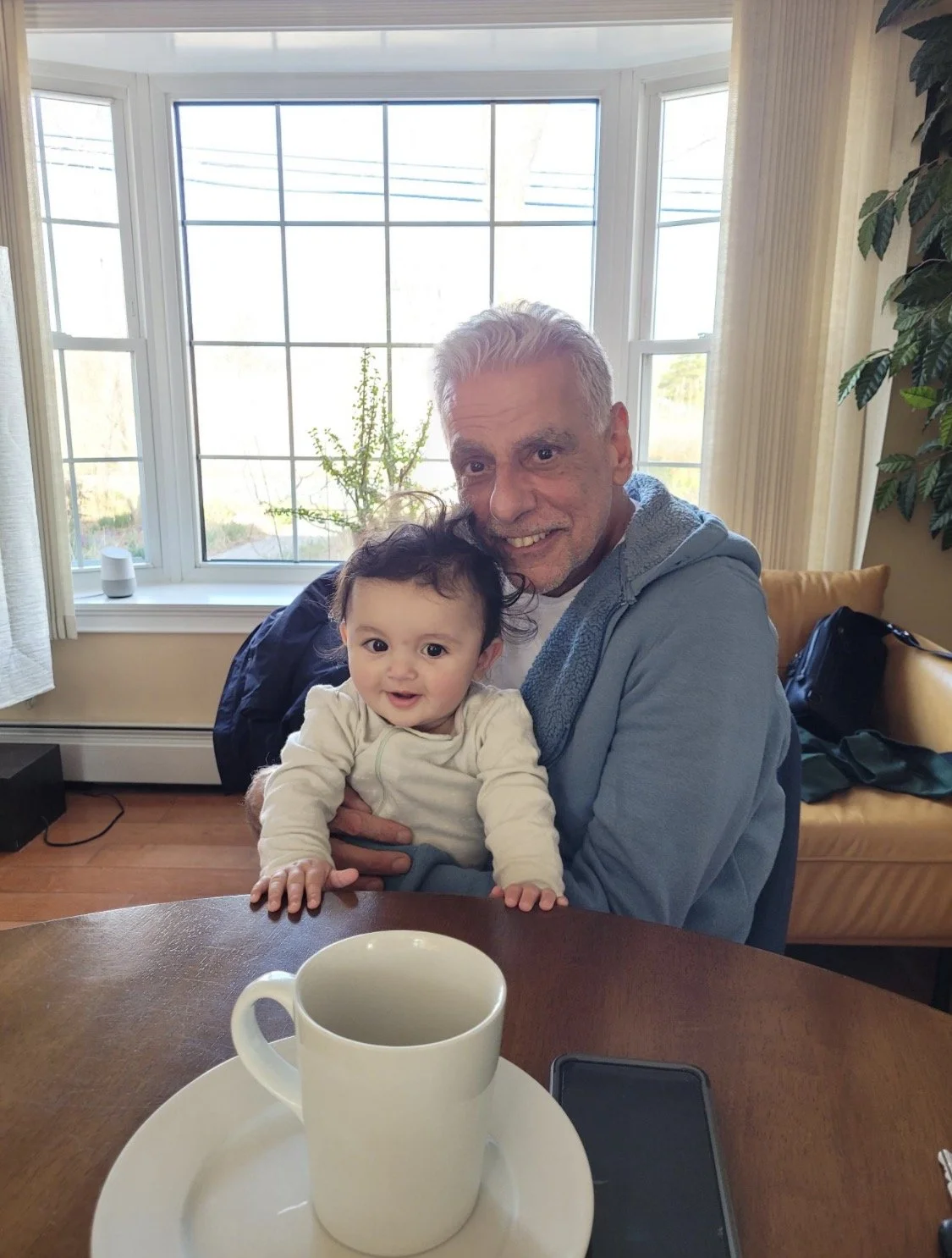 An elderly man with gray hair smiling and holding a young child with curly dark hair and big eyes, sitting at a dining table with a white coffee mug in the foreground, in a room with large windows and natural light.