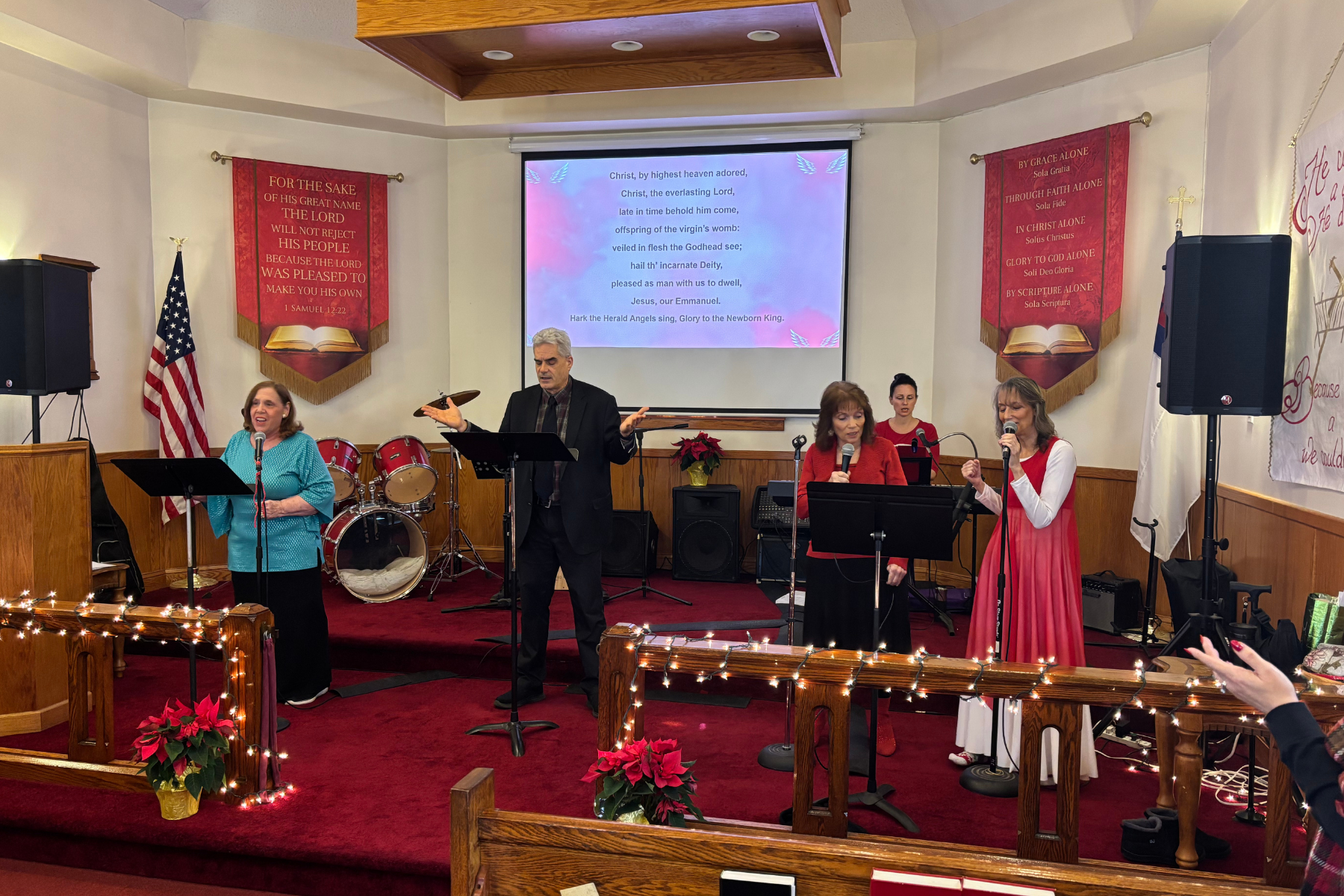 A church stage decorated for Christmas with a group of people singing and playing music. There are three women and one man, with a drum set, speakers, and hymn display. The church features red banners with religious text, an American flag, and poinsettia plants with Christmas lights lining the front.