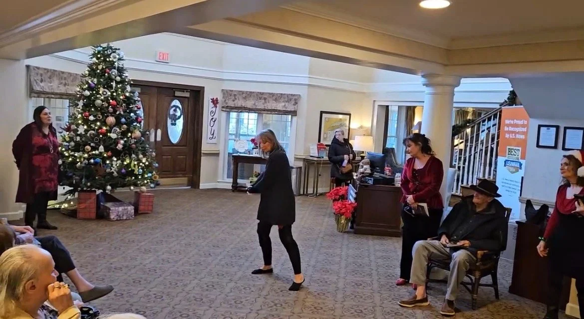 A Christmas scene in a warmly decorated lobby with a decorated tree, presents, and people gathered around, some standing and some seated.