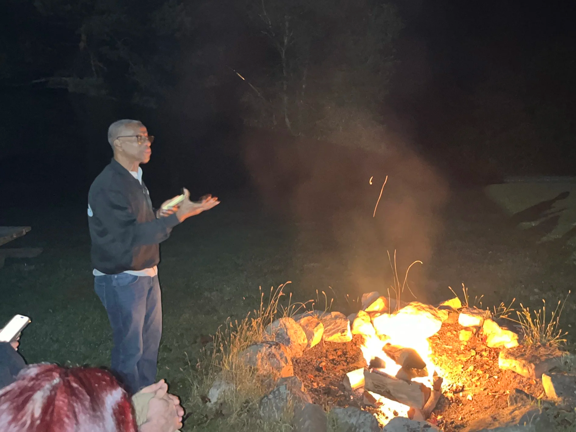A man standing near a campfire at night, holding a book and explaining, with another person partially visible sitting nearby and taking a photo.