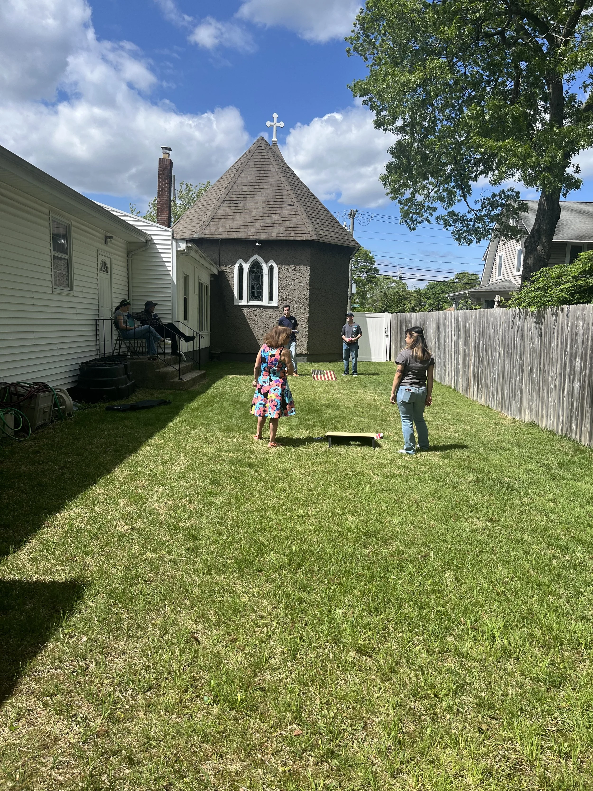 People gathered in a backyard with a church building in the background. The sky is partly cloudy, and there is a grassy lawn with some shaded areas and a wooden fence on the right.