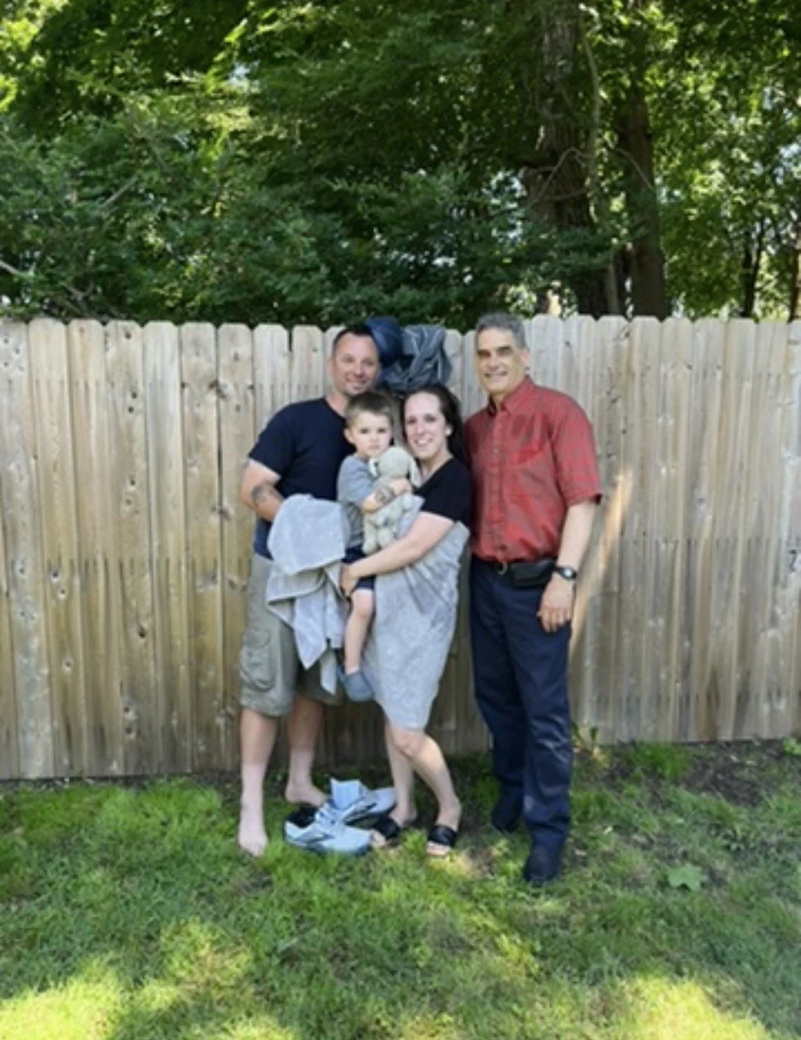 Four people, including a child, standing and smiling in front of a wooden fence with greenery behind it.