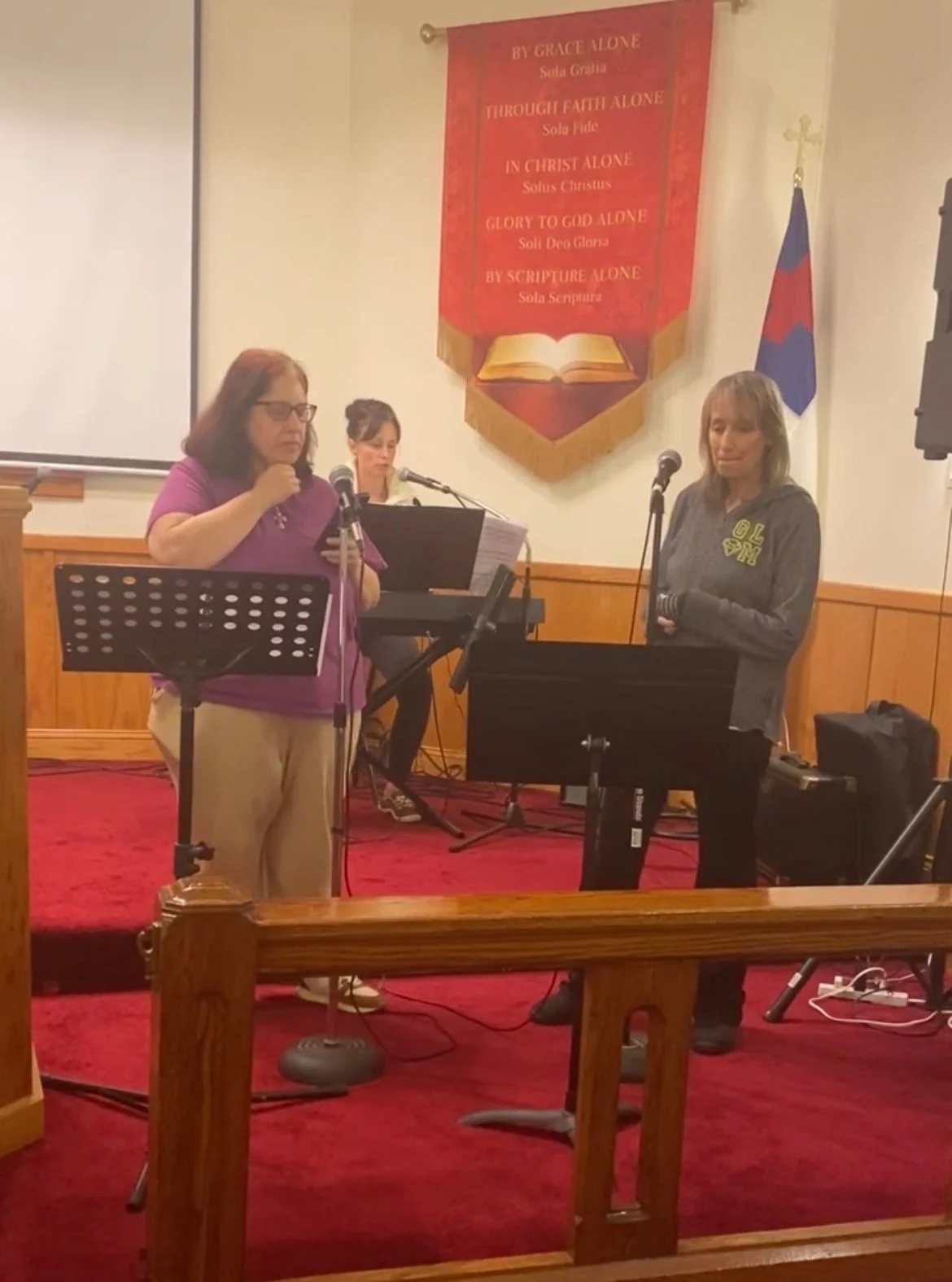 Three women standing on a stage, singing or speaking into microphones, with a woman playing a piano or keyboard behind them. There is a banner on the wall with religious text and an open book, and a red, white, and blue flag to the right of the banner.