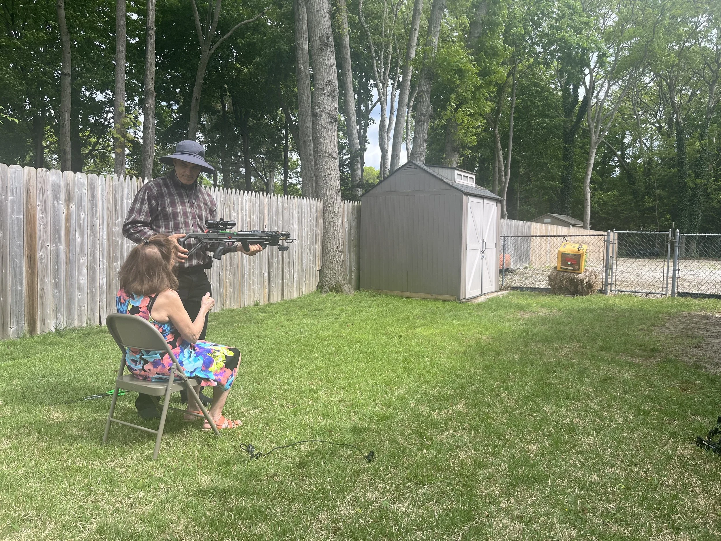 A woman sitting in a folding chair holding a rifle, with an older woman standing behind her pointing at it, in a backyard with a wooden fence, trees, a gray shed, and a chain-link fence in the background.