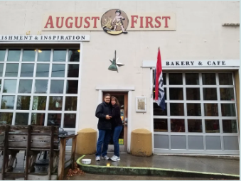 A couple standing in front of a bakery and cafe called August First, with a large window and a door, a flag, and some outdoor seating.