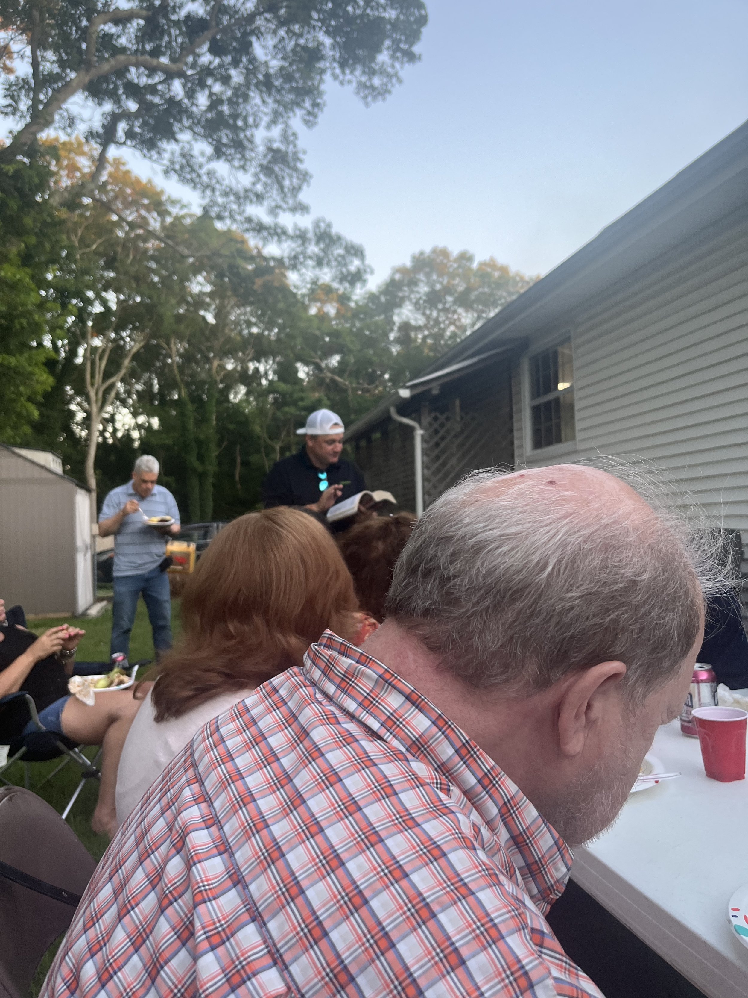 People gathered outdoors for a social event, sitting at a table with food and drinks, with some individuals reading or speaking, and others eating, in a backyard with trees and a house in the background during sunset.