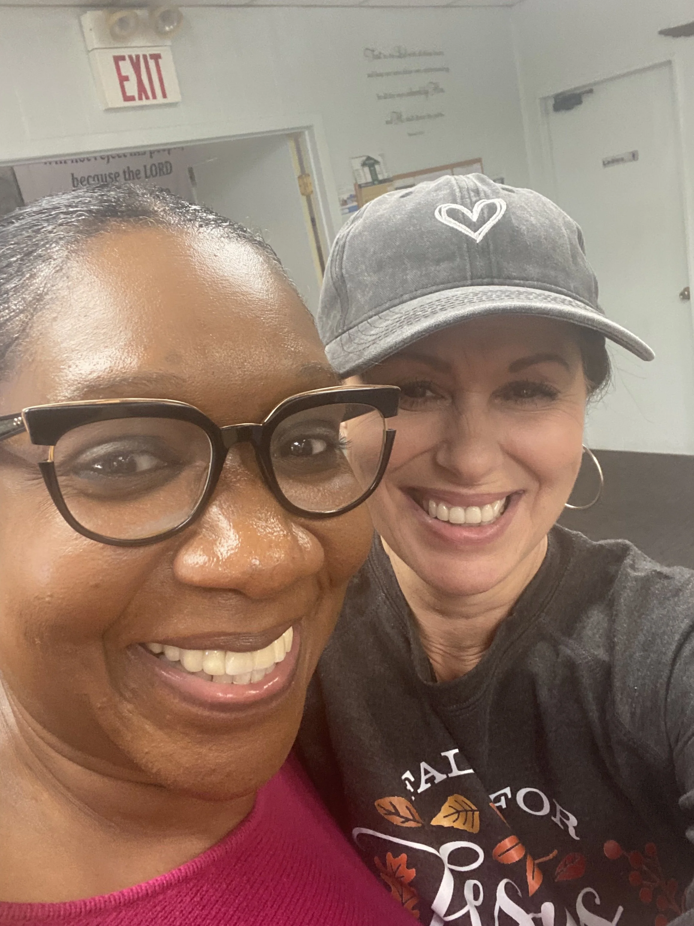 Two women smiling for a selfie indoors, one wearing glasses and a pink top, the other wearing a gray cap and a dark T-shirt with autumn leaves printed on it.