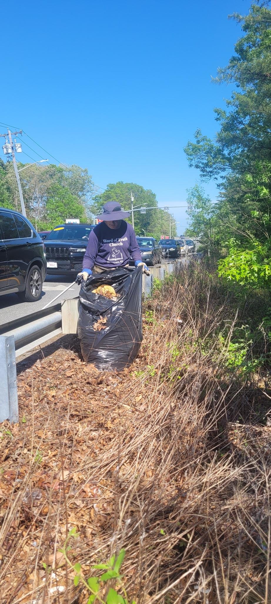 A person wearing a wide-brimmed hat and gloves gathers trash into a large black garbage bag beside a busy road with cars and traffic lights, surrounded by dry leaves and bushes under a bright blue sky.