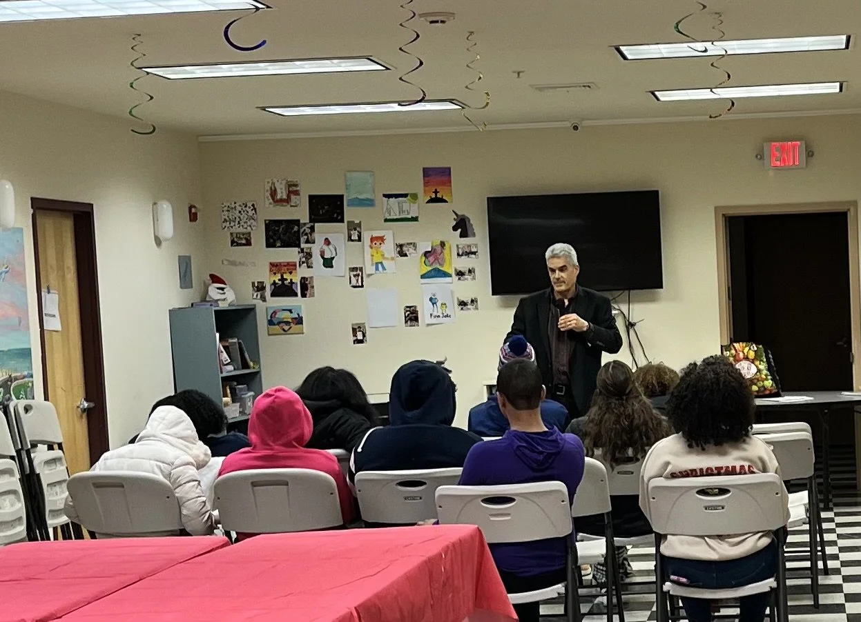A man giving a presentation to a group of children seated in a classroom with colorful artwork on the walls and decorations hanging from the ceiling.