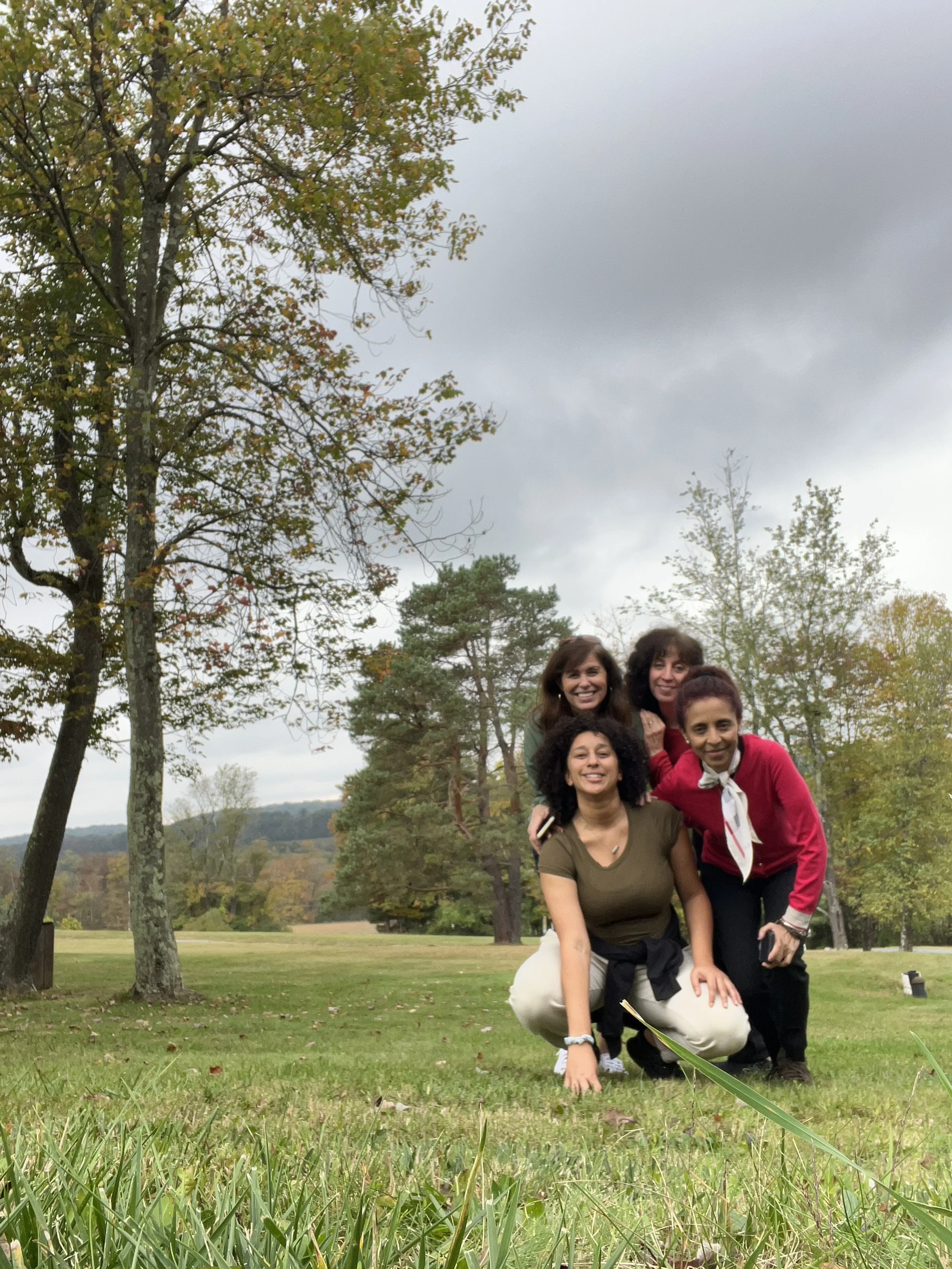 Four women smiling outdoors in a park with trees and cloudy sky, two women crouching and two standing behind them.