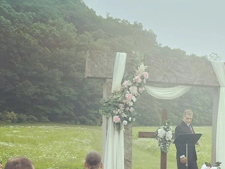Outdoor wedding ceremony with a decorated altar and a man in a suit reading from a book, set against a lush green landscape with trees.